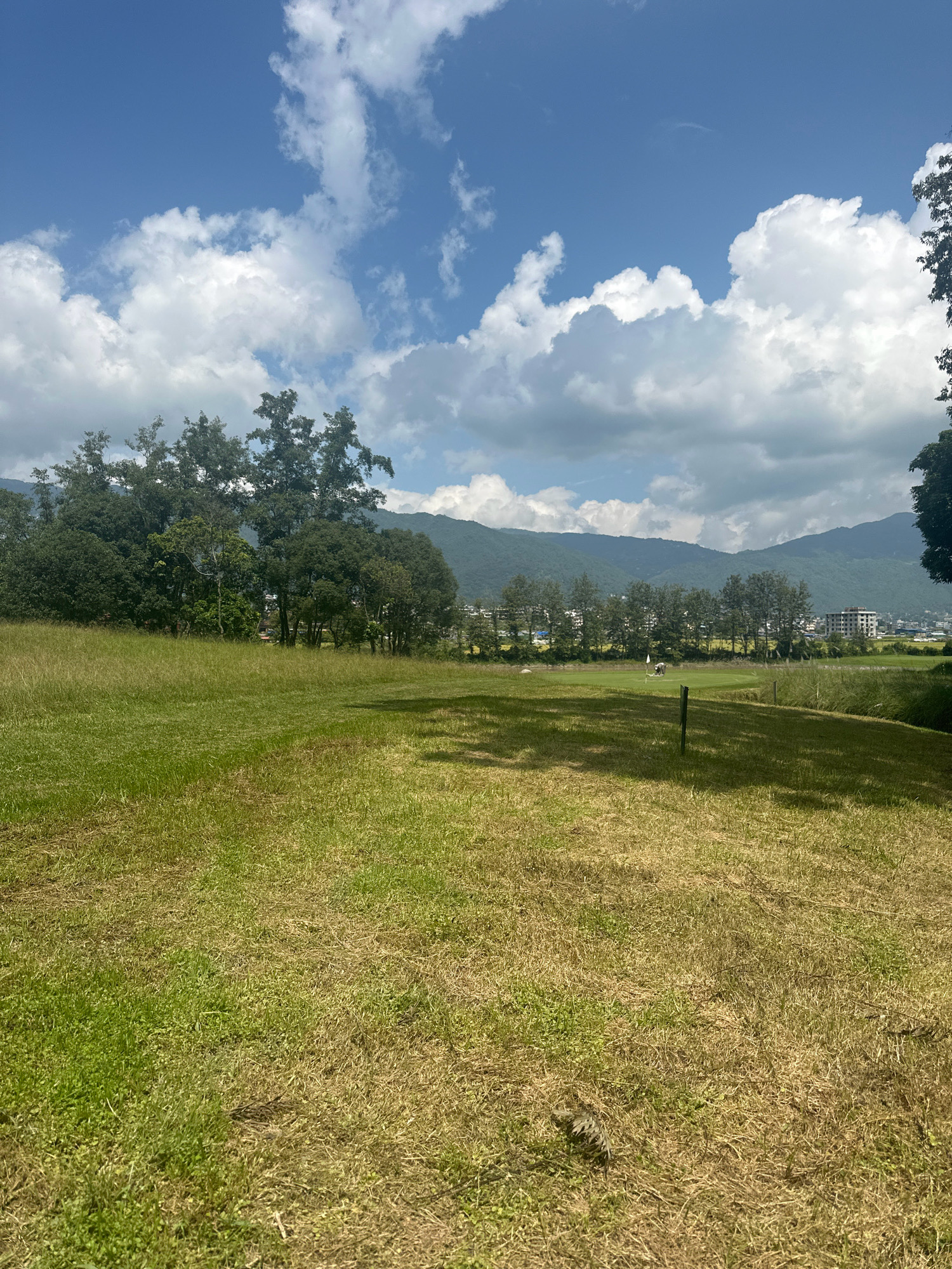 This image depicts a wide, grassy field with a row of trees in the middle distance. Beyond the trees, a town is visible nestled against rolling hills. The sky is a vibrant blue, dotted with fluffy white clouds. In the foreground, the grass is a mix of green and dry, yellowed patches. A solitary wooden post stands in the field. In the middle distance, on a manicured green area, two individuals appear to be playing golf near a flag. A building complex is situated in the background. The overall setting suggests a rural or semi-urban environment in Gokarneshwar, Nepal, likely a golf course or park.