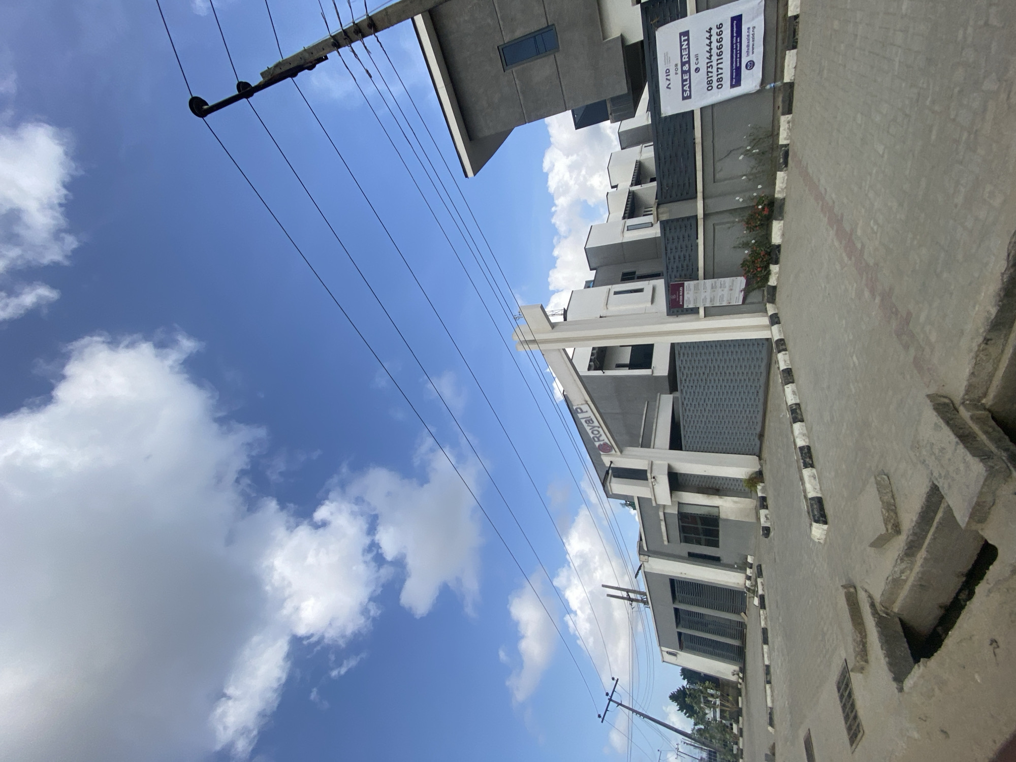 The image depicts a street scene in Lekki, Nigeria, characterized by modern buildings under a partly cloudy blue sky. Power lines are strung across the sky. One of the buildings has a sign with "Royal P" visible. Additionally, a real estate sign advertises properties for sale or rent, displaying the phone numbers "08173144444" and "08171166666." The street is concrete.