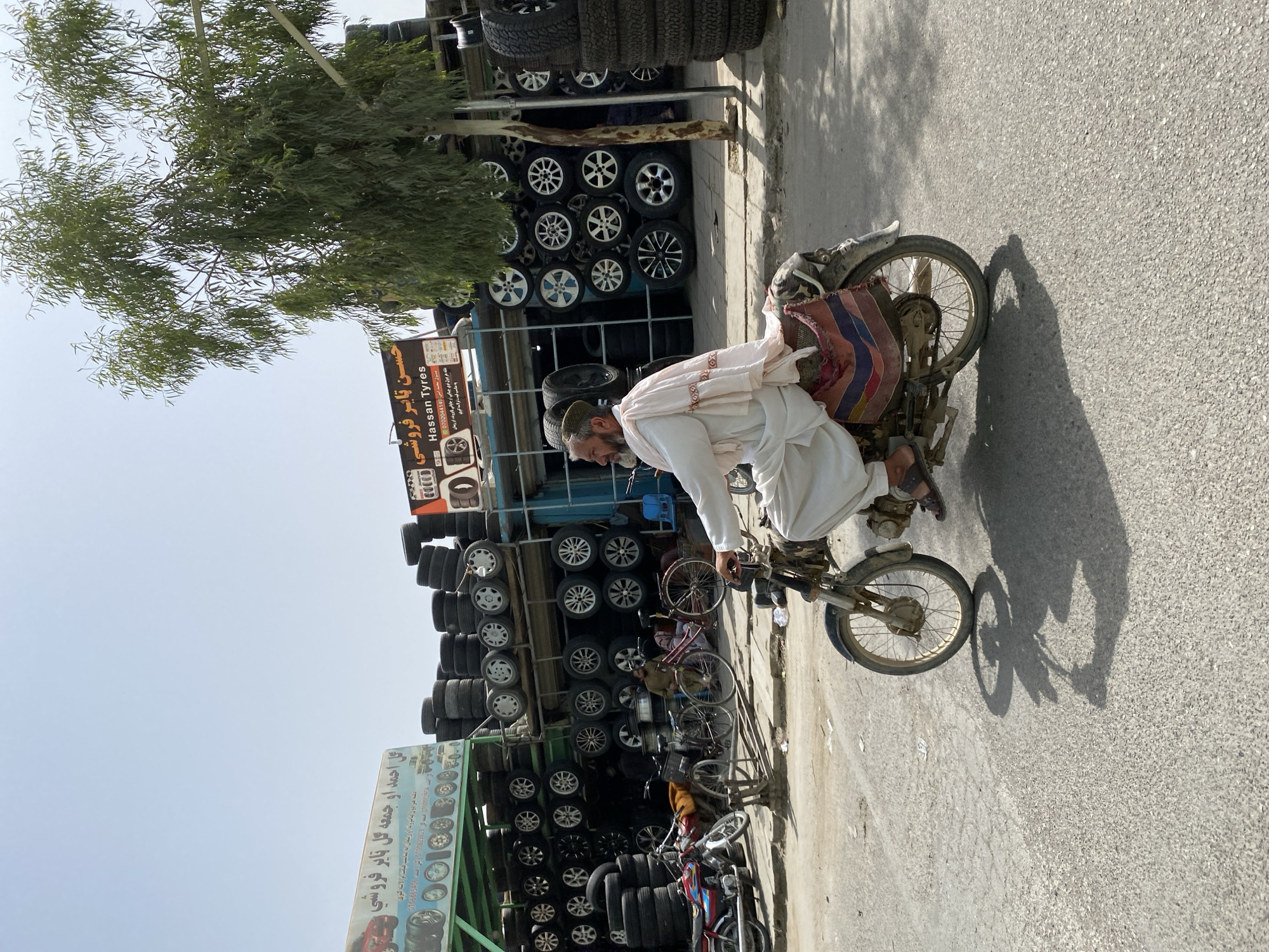 In Kandahar, Afghanistan, a man rides a motorcycle on a paved road. The man is wearing a white shalwar kameez and a green cap. He has a light-colored shawl draped over his shoulders. The motorcycle is old and appears to be a dirt bike. Behind the man, a tire shop is visible, with stacks of tires and car rims displayed on shelves. A sign above the shop reads "Hassan Tyres" in English and Arabic. Several bicycles and other motorcycles are parked near the shop. A large tree with green leaves stands to the right of the frame, casting a shadow on the road. The sky is clear and blue, indicating a sunny day. The scene suggests a typical street view in Kandahar, where commerce and transportation intersect.