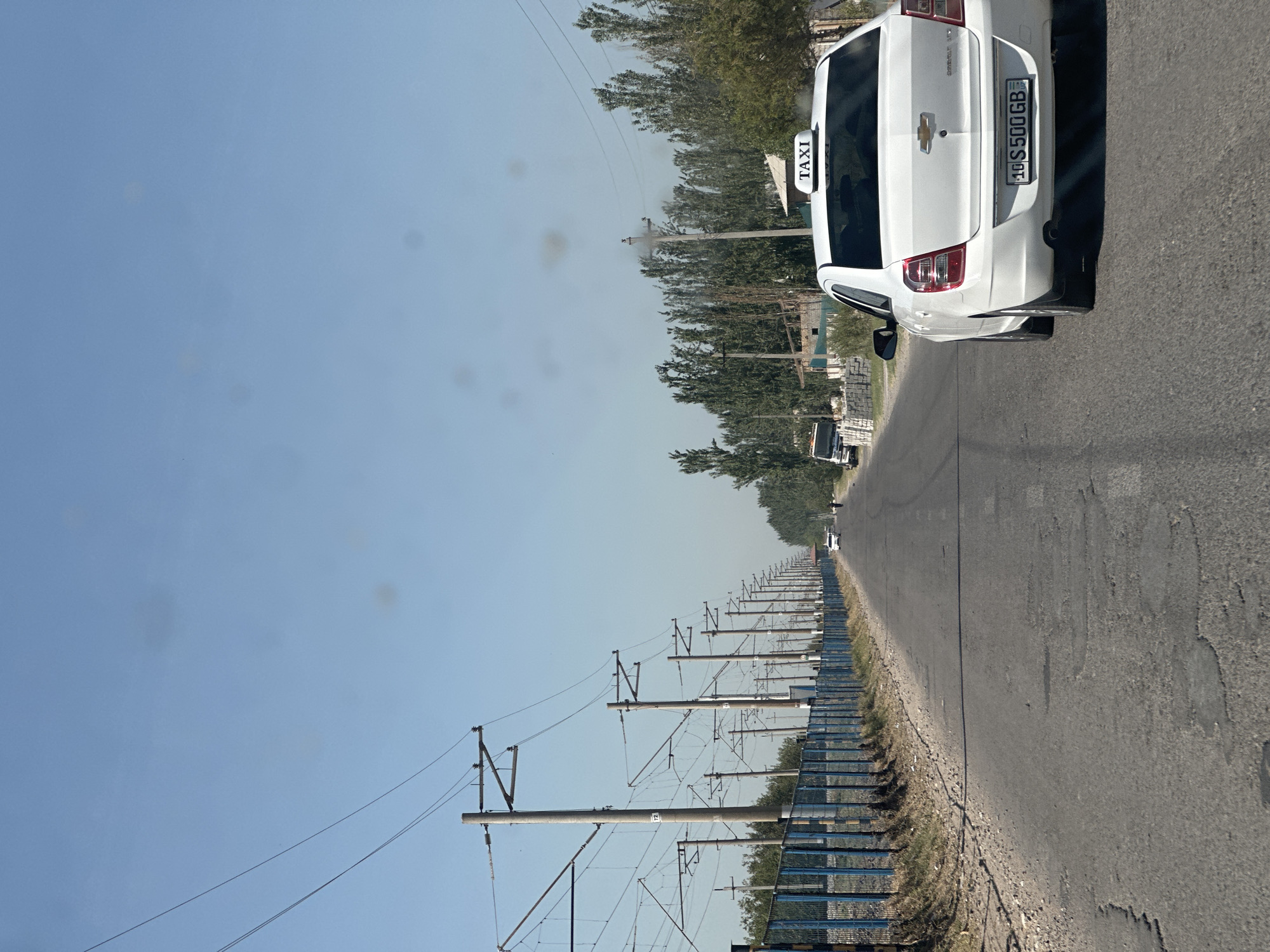 The image shows a straight asphalt road in Yallama, Uzbekistan, lined with power poles and overhead wires on the left side. A blue metal fence runs along the side of the road, adjacent to a patch of dry grass and dirt. On the right side of the road, there are numerous trees and some low-rise buildings. A white Chevrolet Cobalt taxi with a "TAXI" sign on its roof is in the foreground, driving on the road. Its license plate is visible and reads "10 S 500 GB". Further down the road, a few other vehicles and a person are visible in the distance. The sky is clear and blue.