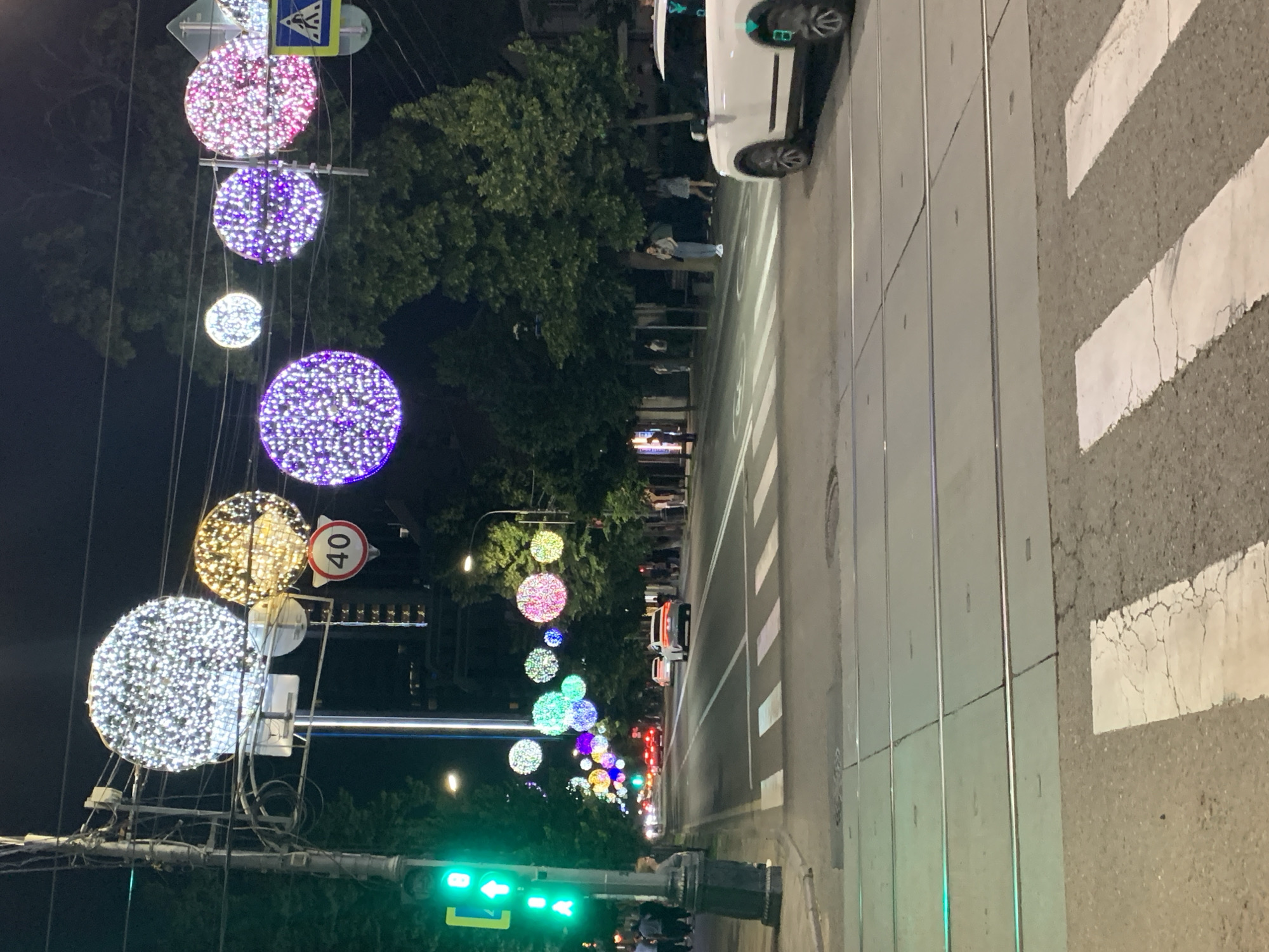 An exterior night shot captures a street scene with overhead decorative lights and cars. The lights, suspended by wires, are large, illuminated orbs in various colors, including white, yellow, pink, and purple. Traffic signals, a speed limit sign indicating 40, and a pedestrian crossing sign are also visible. The street is paved, with tram tracks running across it. Several cars are on the road, with a white SUV prominently featured in the foreground on the right. Pedestrians are present on the sidewalks and near the street. Trees line the street, their leaves dark against the night sky. The overall impression is of a well-lit urban environment at night, possibly decorated for an event or holiday.