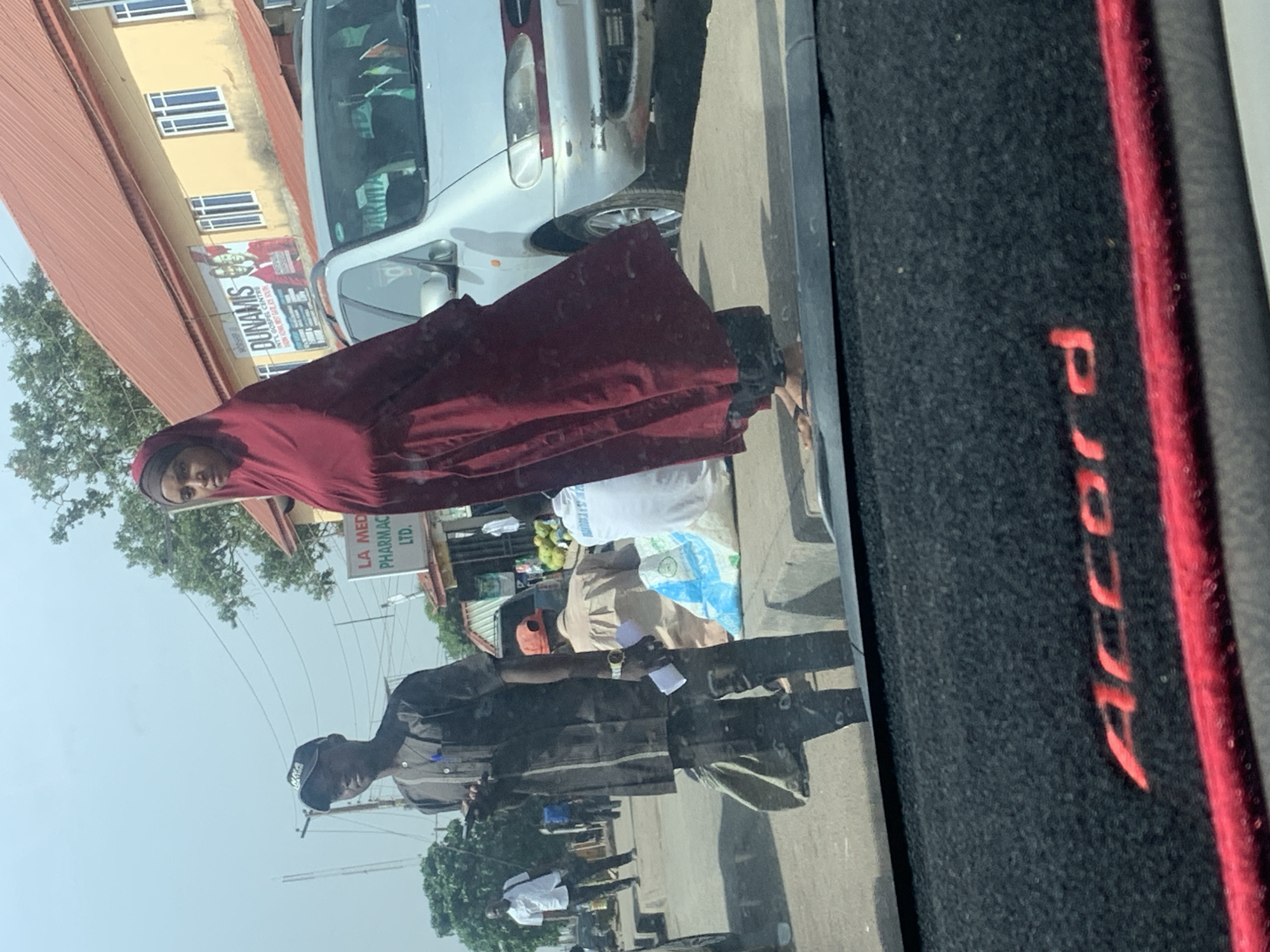 This image captures a street scene in Jos, Nigeria, taken from inside a vehicle, evidenced by the "Accord" lettering visible on the car's interior trim in the foreground. The photo is oriented vertically but depicts a horizontally unfolding street view.
In the mid-ground, a woman wearing a dark red hijab or dress stands on the pavement, looking directly towards the camera with a neutral expression. Behind her, a silver or light-colored car is parked or moving slowly. Further down the street, various individuals are visible; one man in a dark shirt and cap is seen from the back, and another person in a light top and hat is seated near several bags, possibly goods for sale, including some green fruits.
The background features buildings with a reddish-brown corrugated roof and yellowish painted walls. A prominent billboard-like sign advertises "DUNAMIS INT'L GOSPEL CENTRE," with additional text mentioning "WORSHIP" and "DADIN KOWA NRST GATE KOS SOUTH." Another sign clearly reads "LA MED PHARMAC LTD." Trees are scattered throughout the scene, suggesting a semi-urban or commercial area. The bright, clear sky indicates daytime with sunny weather. The overall scene conveys a typical busy street environment in an urban setting.