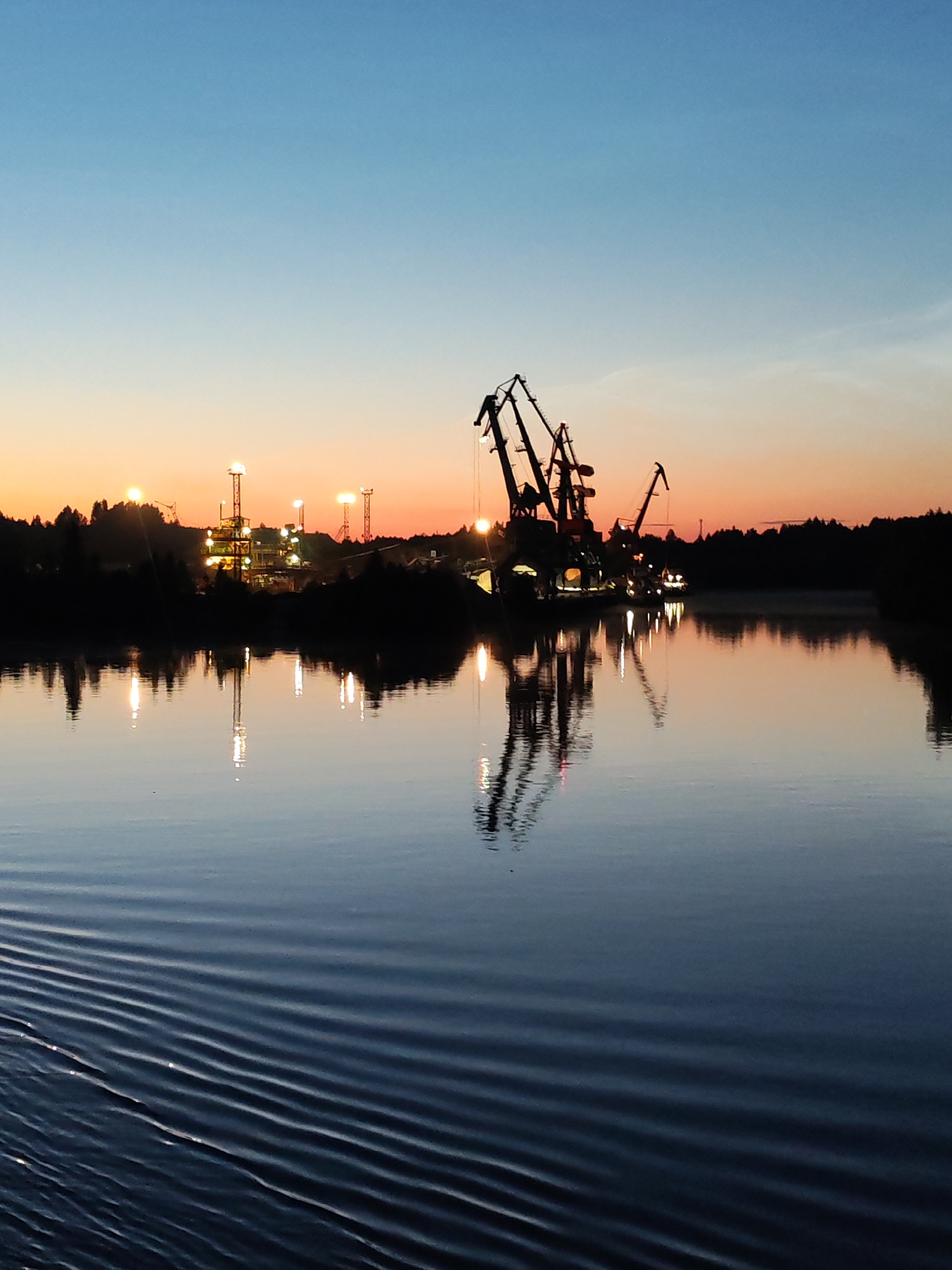 At dusk, a body of water reflects the silhouettes of industrial cranes and lights against a gradient sky transitioning from deep blue to orange. The water exhibits a pattern of ripples on the left side, suggesting movement. The distant shoreline is lined with trees, also silhouetted. The industrial structures, likely part of a port or mining operation, are illuminated by several tall, bright lights. This scene is located near Aleksandrovskoe, Russia.