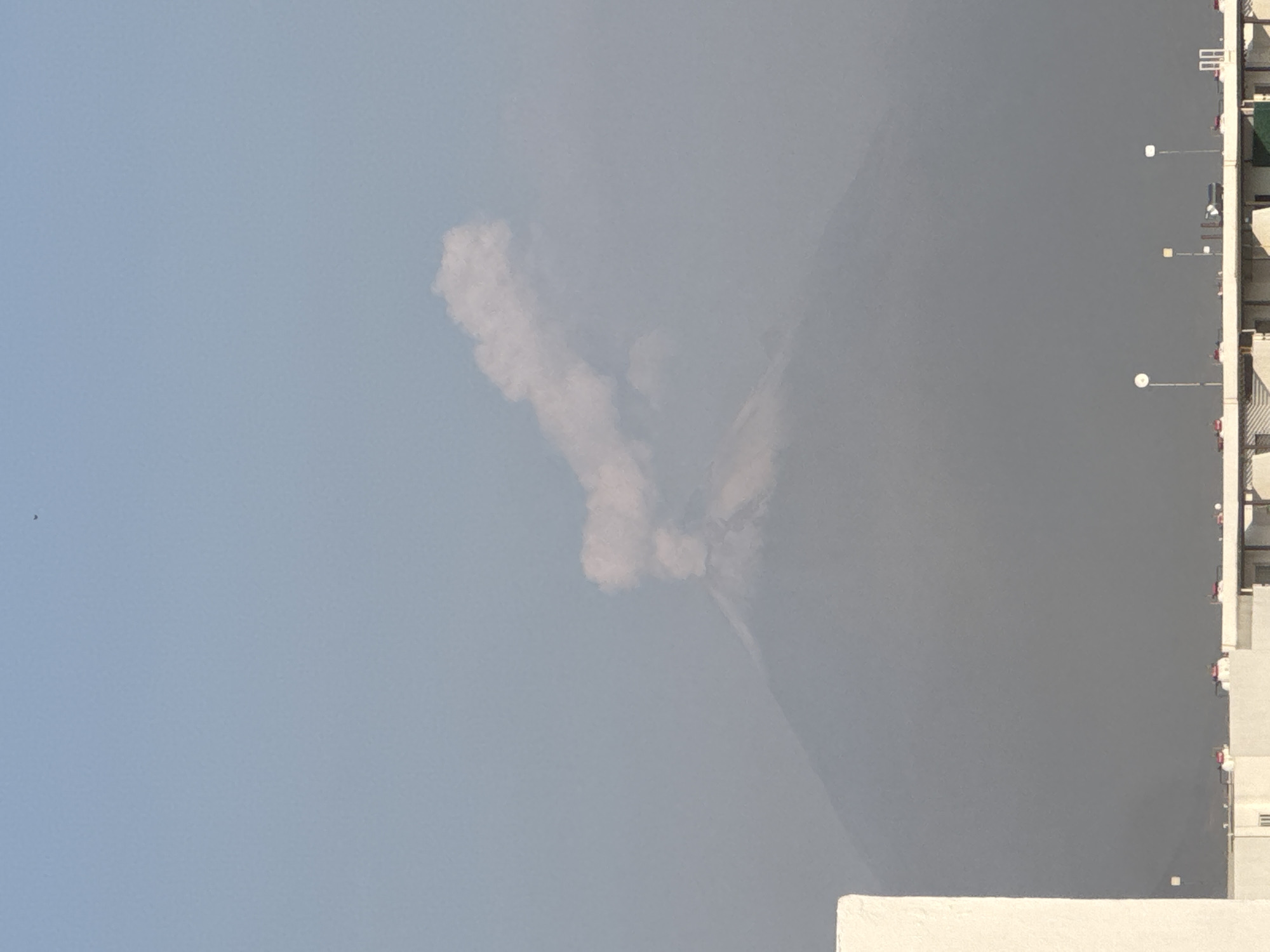 The image shows a daytime view in San Francisco Ocotlán, Mexico. The sky is light blue with a small black speck that may be a bird. A large plume of light gray smoke rises from a gray landmass, possibly a mountain or hill. A tall, white building with multiple stories and exterior lights is visible along the right edge of the image. Part of a white wall is also visible in the bottom right corner.