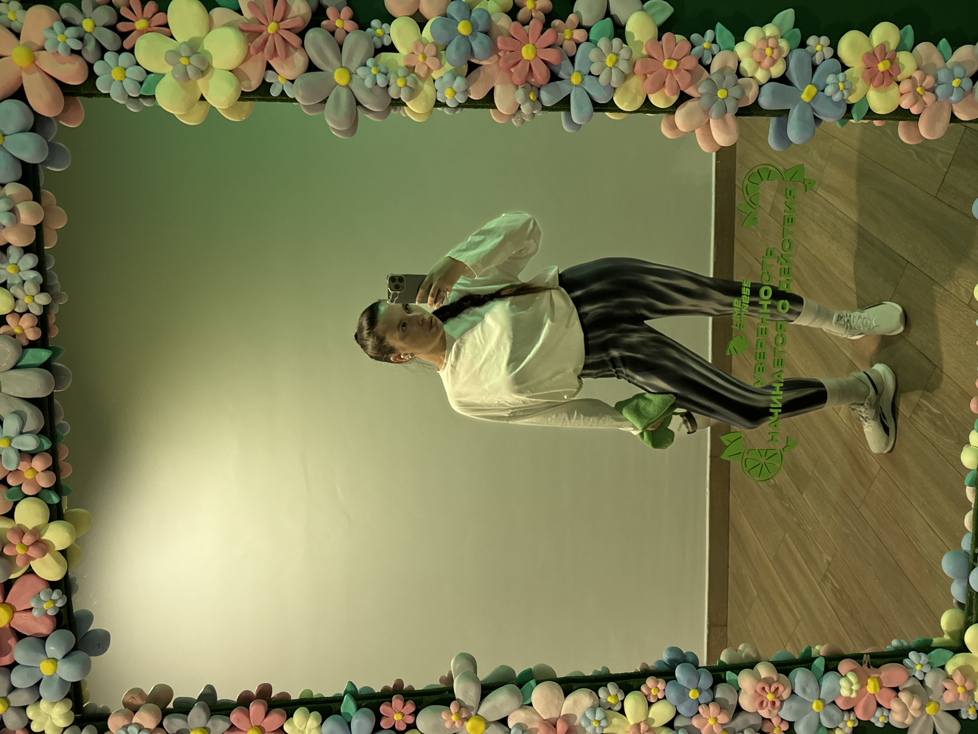 An indoor mirror selfie captures a young woman standing in front of a mirror with a vibrant, floral frame. The image, rotated 90 degrees clockwise, shows the woman sideways, extending her right arm to hold a dark smartphone and take a picture of her reflection. She wears a white long-sleeved shirt, dark and light striped leggings, white socks, and white sneakers. Her dark hair is styled in a braid, and she holds a small, folded green object in her left hand.
The mirror's frame is distinctly decorated with numerous soft, pastel-colored, three-dimensional flowers in shades of pink, blue, yellow, light green, and light gray, creating a whimsical border around the reflection.
Reflected in the mirror is a light-colored wall and a light-toned wooden floor. On the wooden floor, visible on the right side of the reflection, is green Cyrillic text and a logo. The mirrored text reads "УВЕРЕННОСТЬ ДЕЙСТВИЯ НАЧИНАЕТСЯ," which translates to "Confidence of action begins." Above this, a small logo resembling a lime or citrus fruit is present, along with text that appears to say "LIME FITNESS."
The setting is an interior space, likely a commercial establishment such as a fitness center or a creatively decorated area, suggested by the fitness branding and the activewear worn by the woman. The lighting is soft and diffused, indicating an indoor environment. The location context is Vniissok, Russia.