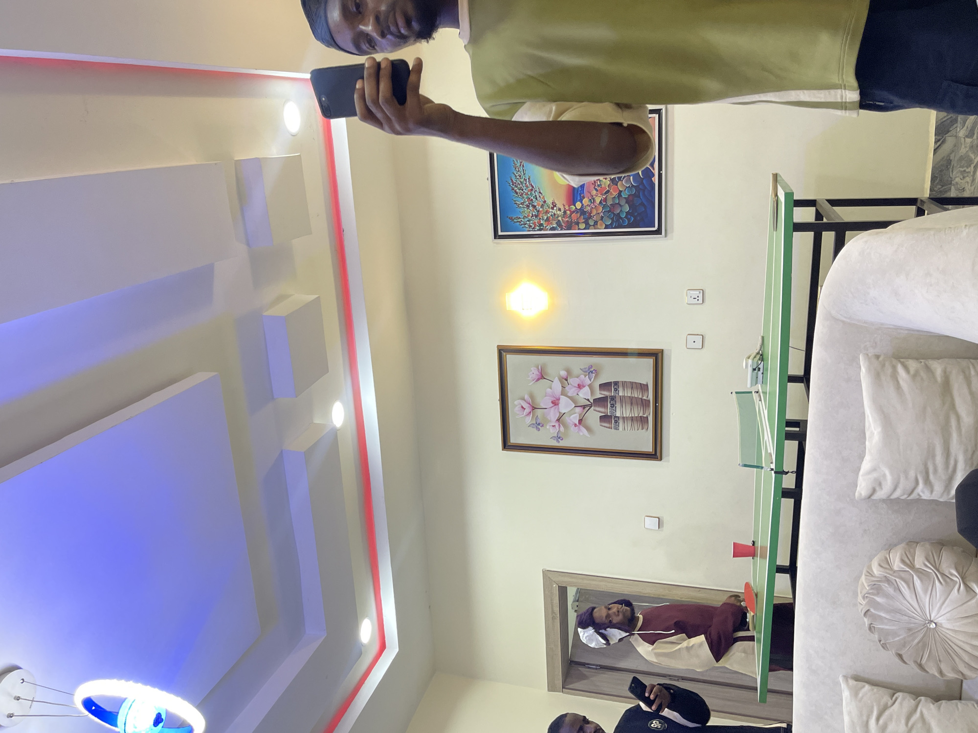 This portrait-oriented image captures an indoor scene, likely a living room or recreational area in Gwarinpa, Nigeria. The room features a modern aesthetic with a false ceiling showcasing geometric patterns and various lighting fixtures, including recessed white lights, warm yellow wall sconces, and a distinctive blue and white circular chandelier. The walls are painted a light, neutral color.
In the left-center of the frame, a man with a beard and a dark head covering stands, looking directly at the camera with a slight smile. He is wearing a light green short-sleeved top and dark trousers, and holds a black smartphone in both hands.
Towards the right side of the room, a light-colored sofa with several throw pillows (one white, one round beige) is visible. In front of the sofa, a compact green table tennis table is set up with a net, a red cup, and a paddle, suggesting a recreational atmosphere.
Two framed artworks adorn the back wall: one colorful, abstract piece above a second painting depicting pink flowers in vases. Below the floral painting, a mirror reflects another man, also with a dark head covering and beard, wearing a maroon and beige long-sleeved top. He appears to be standing near the table tennis table. Partially visible in the lower left corner is another person's head and shoulder, wearing a dark top and holding a smartphone.
The scene is well-lit by artificial lights, making it difficult to determine the specific time of day. No visible text is present in the image. The overall mood appears casual and relaxed, with individuals engaged with their phones and the presence of a game table.