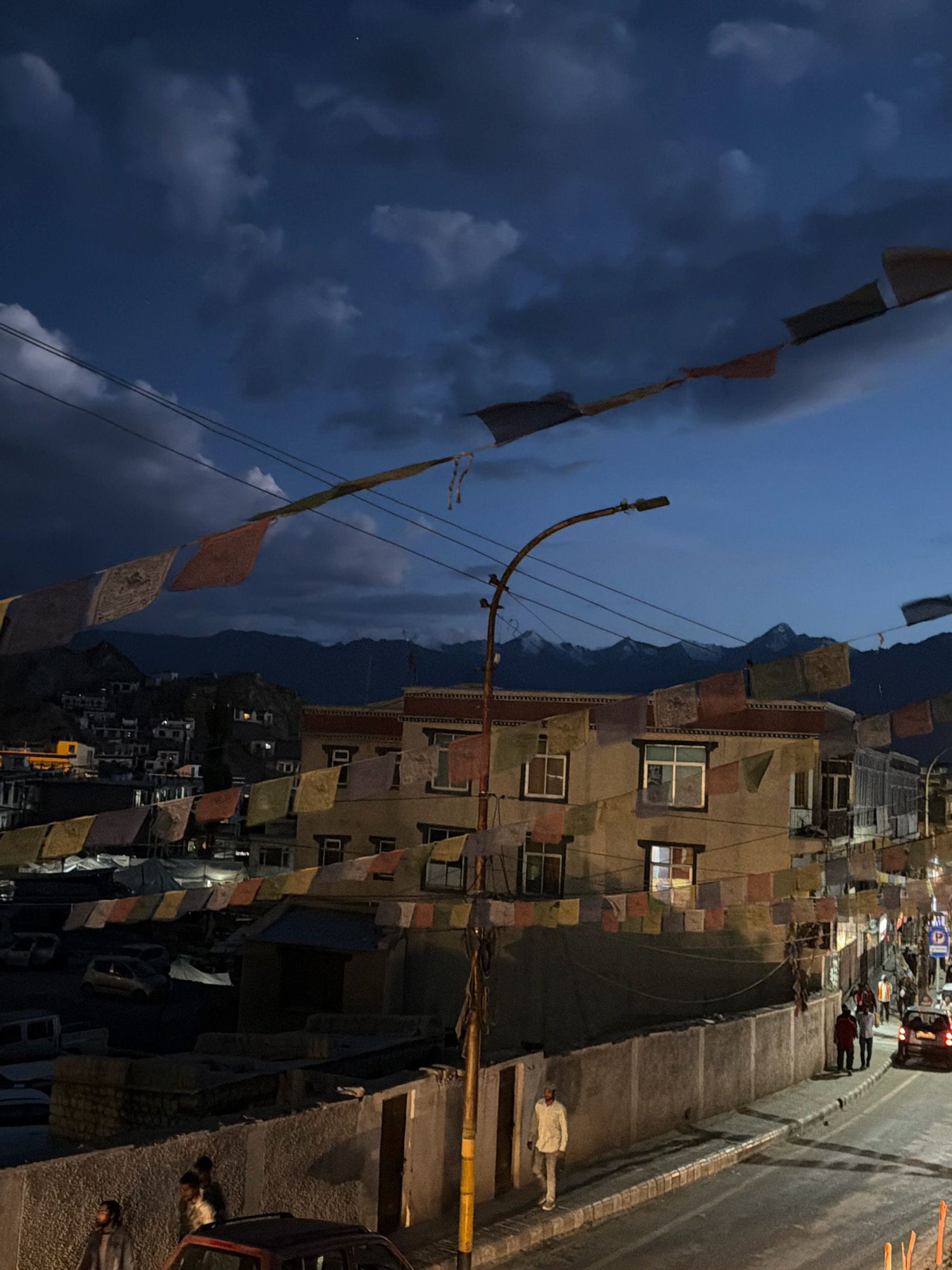The image captures an evening scene in Leh, India, likely on Leh-Manali Highway, given the mountainous backdrop and Buddhist prayer flags, common in the region. The sky is a deep twilight blue with dark, dramatic clouds, and a few stars are visible. Prayer flags, in various colors and patterns, are strung across the scene, some fluttering in the wind.
In the foreground, a street is visible with a paved road and a sidewalk. Several cars are parked or driving on the road. A concrete wall runs along the sidewalk. On the sidewalk, there are a few people, one in a white shirt and light-colored pants walking away from the camera. Further down the street, a group of people are walking, and a car is turning a corner.
Behind the sidewalk and wall, there is a building with multiple stories, adorned with more prayer flags. The building has several windows, some illuminated from within. In the background, a densely populated hillside with houses and lights is visible, set against a backdrop of dark, imposing mountains, their peaks dusted with snow. A street lamp on a pole is also present in the midground, contributing to the evening ambiance. The overall impression is one of a calm, yet vibrant, evening in a mountainous town.