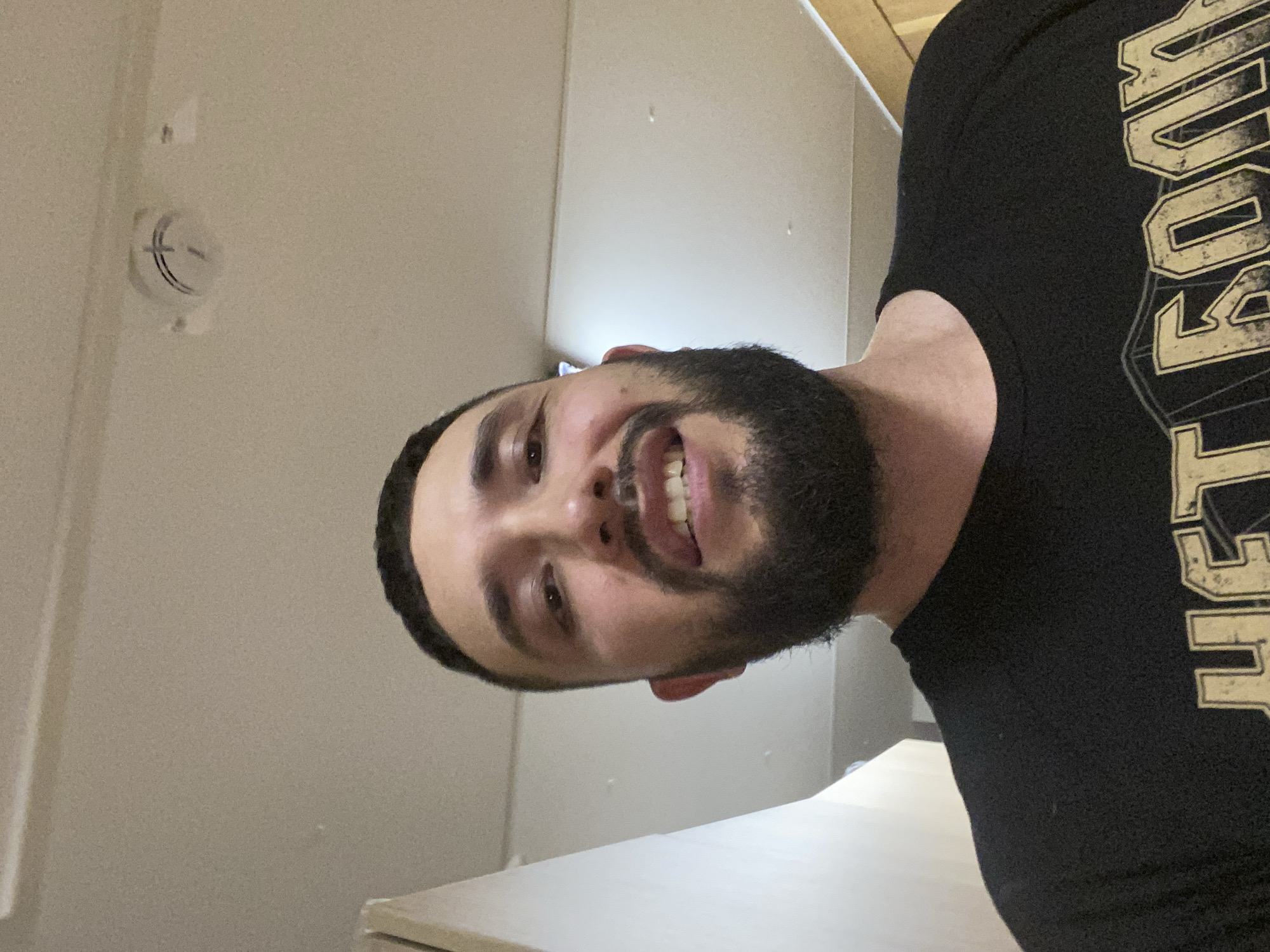 A man with a beard is smiling at the camera. He is wearing a black t-shirt with gold lettering. The background is a light beige wall with a smoke detector. There is also a white surface, possibly a countertop or table, in the foreground. The location is unknown and is not visible in the image. The information requested in the prompt, "location in the city of Koskolovo, Russia", cannot be determined from the image provided.