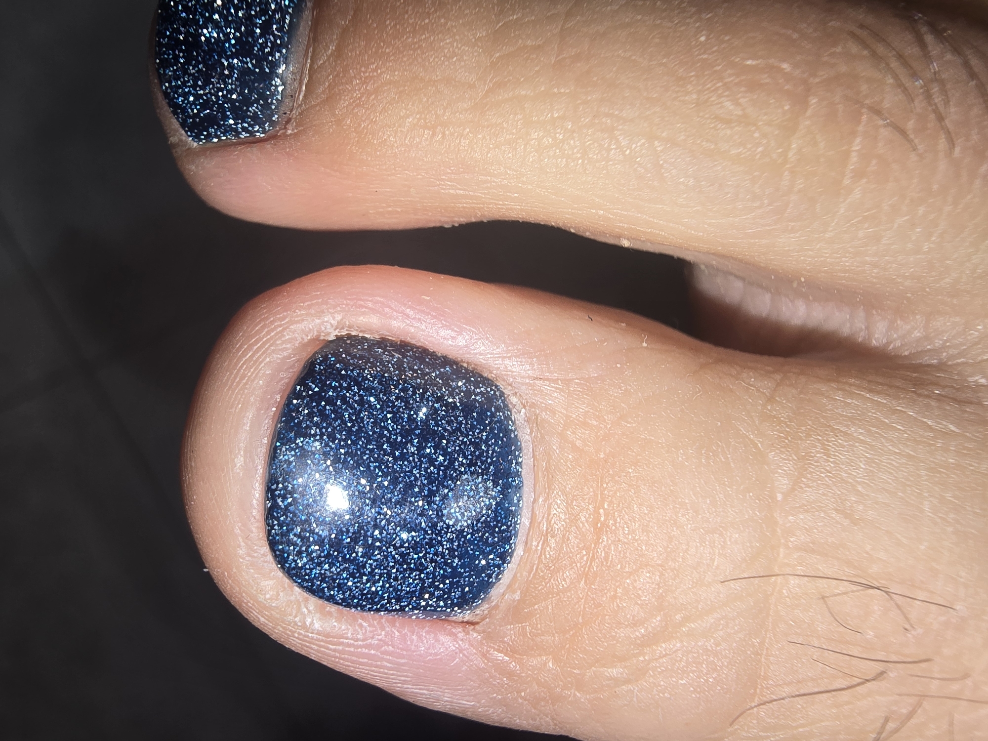 The image is a close-up, low-angle shot of two toes, presumably from a human foot, with fingernails painted in a dark, glittery blue polish. The skin of the toes appears textured with fine lines and has a few dark hairs visible. The lighting creates highlights on the polished nails and casts shadows on the surrounding skin and the background, which is dark and indistinct, suggesting an indoor setting with artificial lighting. There is no visible indication of the location in Ulan-Ude, Russia.