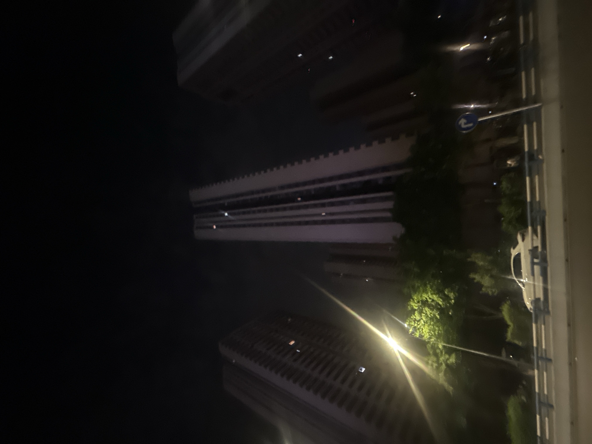 The image captures an urban night scene in the Chongqing High-tech Zone, China, with the camera oriented vertically. Several tall buildings, mostly dark against the night sky, feature illuminated windows, suggesting active occupancy. One prominent building exhibits a distinctive serrated pattern along its top edge.
On the right side of the frame, a road or street is visible, delineated by white and blue guardrails. Lush green foliage of trees is brightly illuminated by an artificial light source, possibly a street light, which creates a prominent lens flare in the lower right portion of the image.
A notable detail is a blue, circular traffic sign with a white arrow pointing right and slightly upwards, indicating a directional instruction, likely a mandatory right turn or curve. A faint, light-colored vehicle can be seen behind the guardrails and trees. The overall setting is dark, emphasizing the artificial light sources and the quiet stillness of the city at night. No people or specific activities are visible.