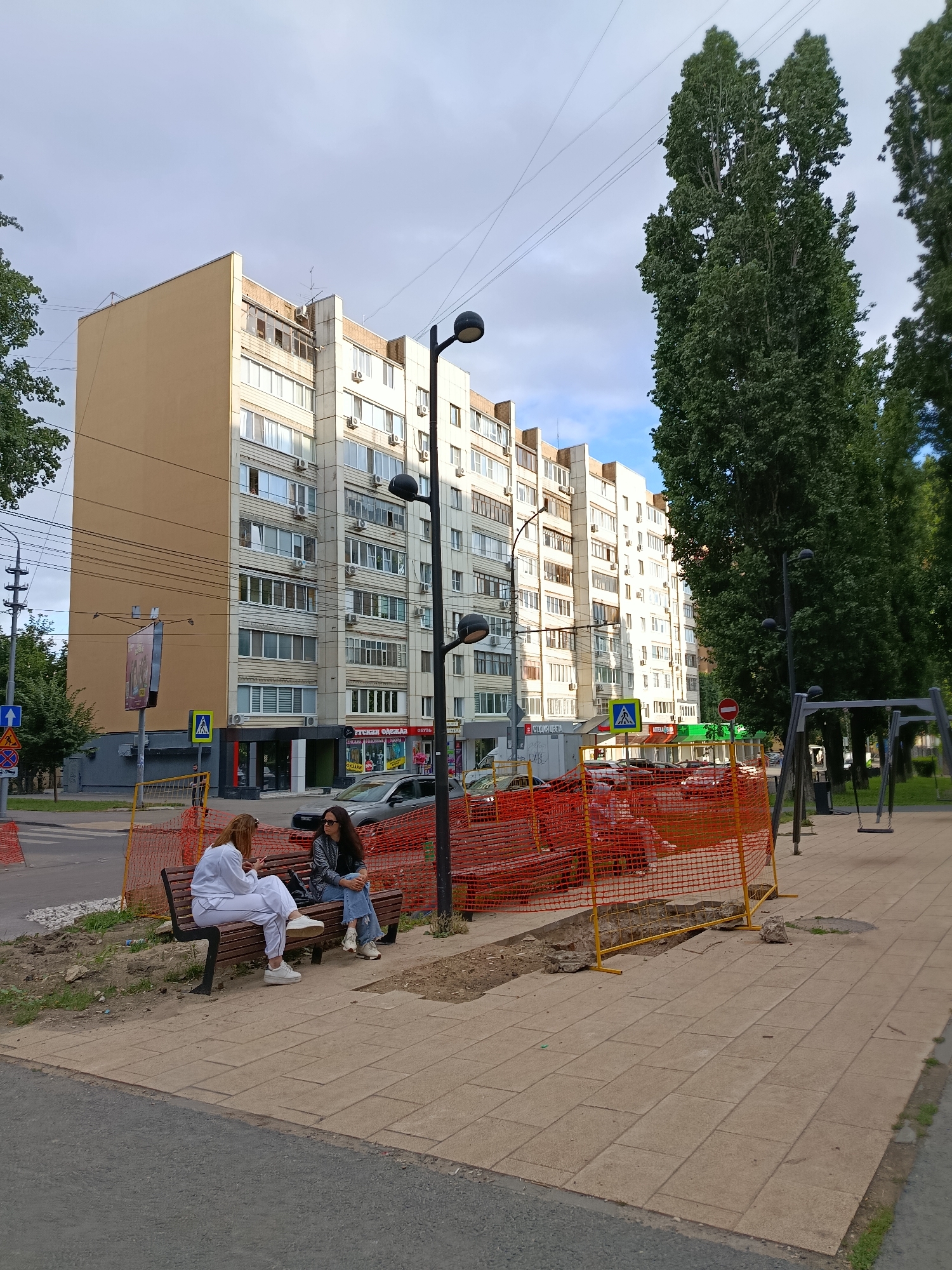 Two women are sitting on a park bench on a paved area next to a construction zone in the city of Saratov, Russia. The woman on the left is wearing white sweatpants and a white jacket, and the woman on the right is wearing ripped blue jeans and a sparkly jacket. Both women are looking down at their phones. The construction zone is enclosed by orange plastic fencing and yellow poles. In the background, there is a tall, light-colored apartment building with many windows. There are also tall green trees on the right side of the image. The sky is partly cloudy.