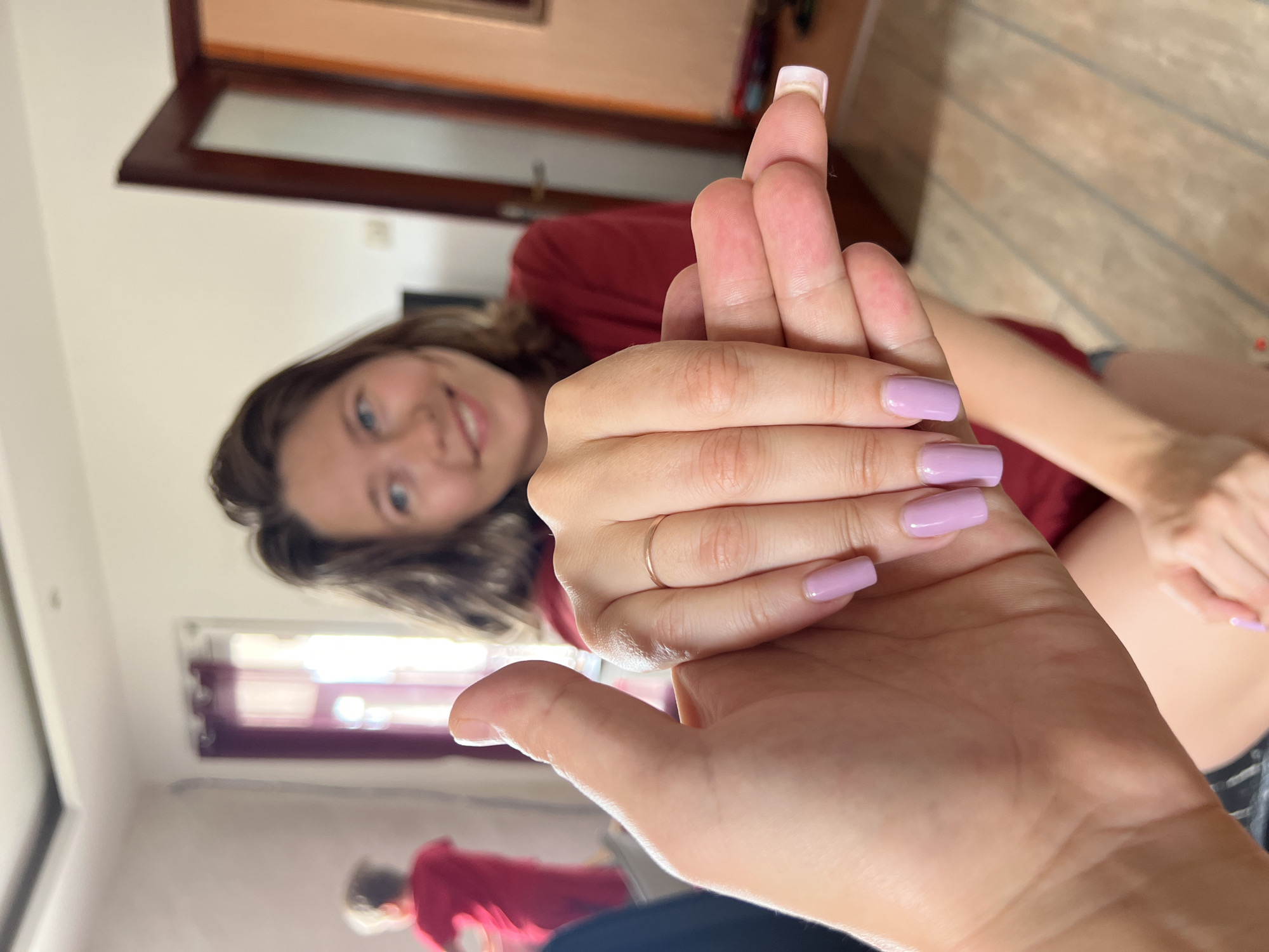 A low-angle, medium shot depicts a young woman holding up her middle finger towards the camera. Her face is visible in the background, showing a slight smile. She has brown hair and is wearing a red shirt. The hand closer to the camera has lilac-colored nail polish and a gold ring on the ring finger. A portion of another person wearing a red shirt is visible near a window in the background, suggesting the possibility of a mirror reflection. The environment appears to be inside a residential space. The location could be Podgorica, Montenegro, but the interior setting provides no visual clues to confirm this.