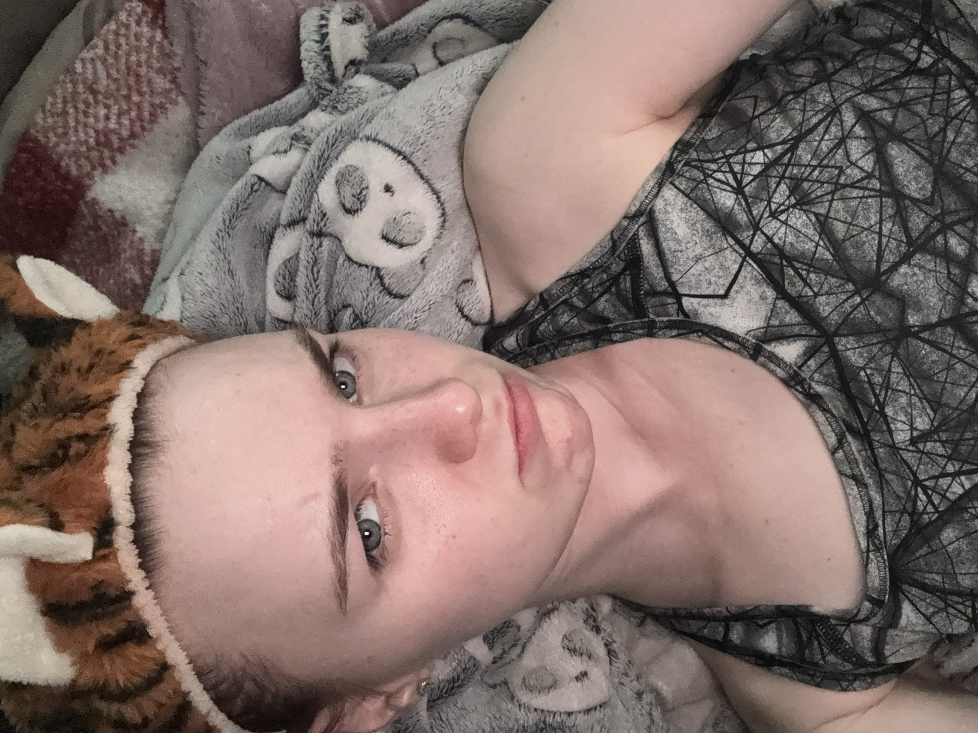 A young woman is lying on a bed, looking towards the camera with a slightly displeased expression. She is wearing a tiger-striped headband with ears and a grey tank top with a geometric pattern. She is covered by a soft, grey blanket with panda faces printed on it, and a red and white plaid blanket is visible behind her. The environment appears to be a bedroom.