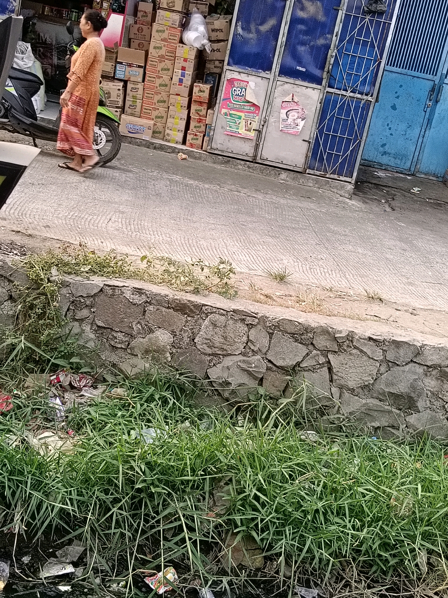 An Indonesian woman walks on a sidewalk in front of a store in Belimbing, Indonesia. She is wearing an orange dress and sandals. The store has stacks of boxes in front of it, and a motorcycle is parked next to the woman. A stone wall is visible behind the sidewalk, with some greenery growing on top of it. The ground in front of the wall is covered with grass and some scattered trash. The scene appears to be an outdoor marketplace or a street in a residential area.