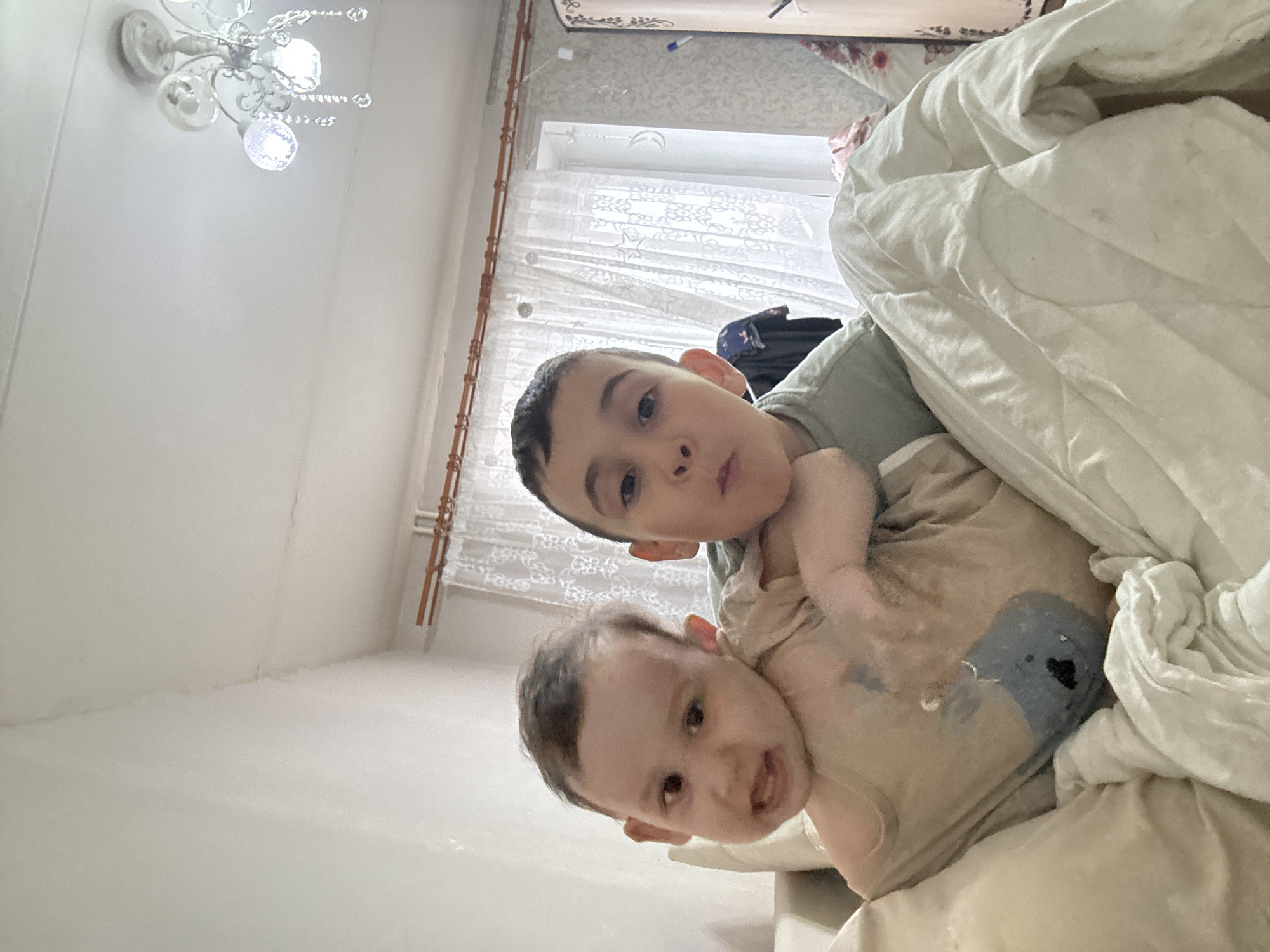 The image shows two children in what appears to be an indoor setting. An older boy with short dark hair and brown eyes is looking at the camera. He is wearing a green shirt. A younger baby is beneath him wearing a light-colored shirt with a blue image on it, also looking toward the camera. Both children seem to be lying on a bed with a white comforter. In the background, a window with white curtains is visible. A chandelier is visible on the ceiling. Based on the background, the location is possibly in an apartment in Chaikovsky, Russia.