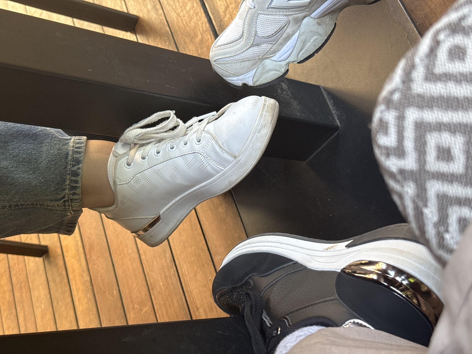 The image shows a close-up of three pairs of feet wearing sneakers, resting on a wooden deck, near a dark metal table leg. One person wears white sneakers with gold accents, another black and gold sneakers, and a third person wears light gray sneakers. The scene appears to be outdoors. The location is not identifiable as Sofia, Bulgaria.