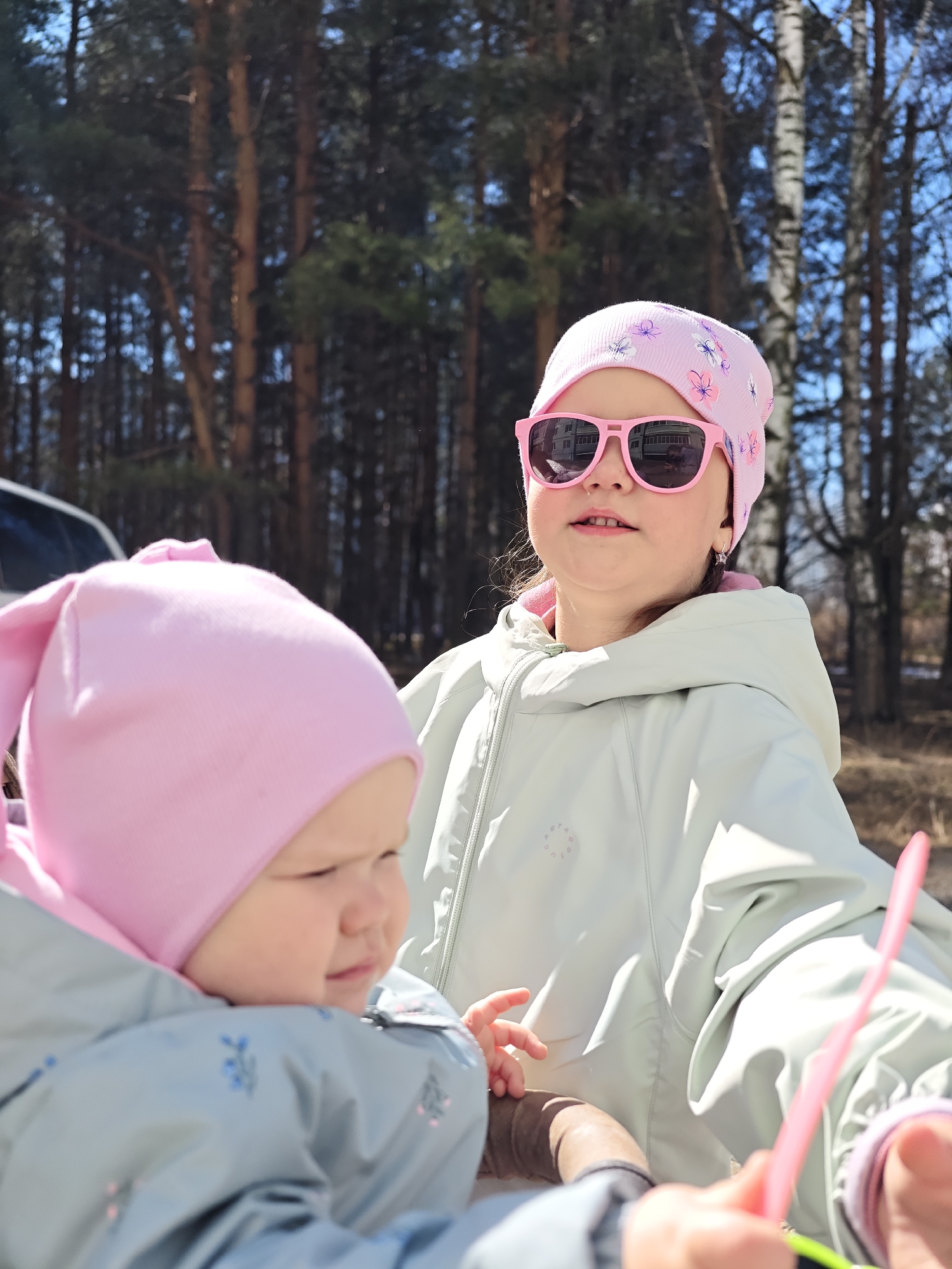 Two young girls are in an outdoor setting, possibly a park or wooded area. The older girl wears a light green jacket, a pink hat with flower patterns, and pink sunglasses. The younger child is dressed in a gray-blue jacket and a pink hat. Trees form the background, with sunlight filtering through the branches. A car is partially visible on the left side of the image. The location appears to be in the city of Chaikovsky, Russia.