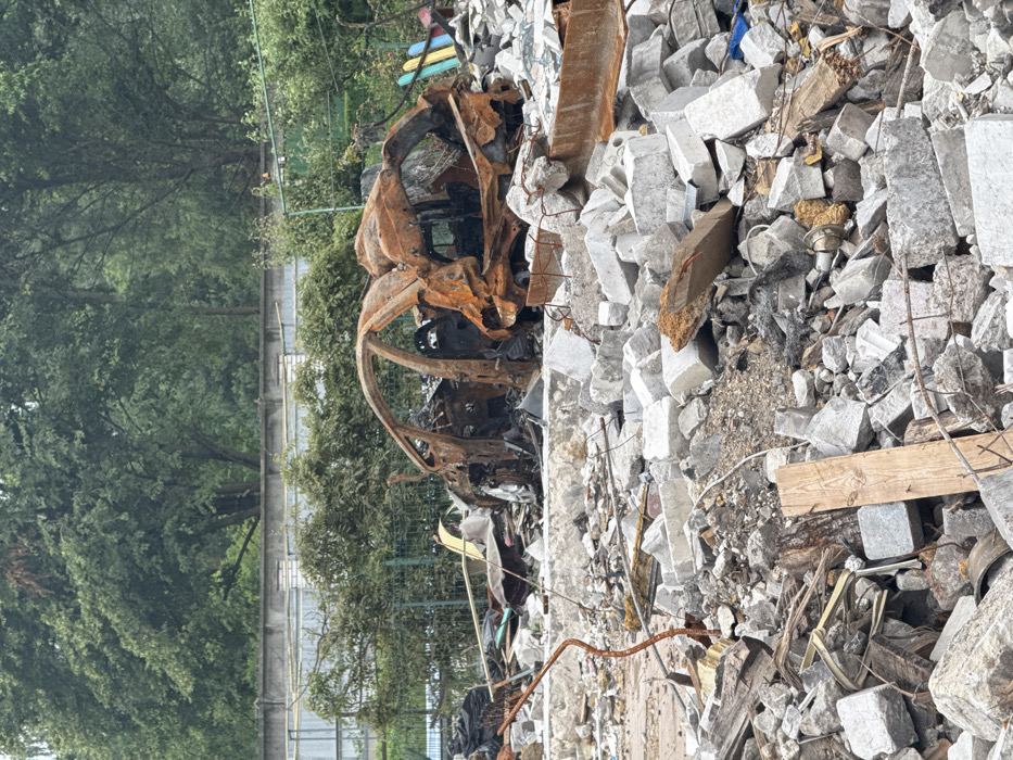 A scene of destruction. A severely burnt-out car sits amidst rubble, consisting of broken concrete blocks and splintered wood. Lush green trees and a fence form a backdrop to the devastation. The car's twisted metal frame is the focal point, indicative of intense heat and impact. The surrounding debris suggests a building collapse or other significant event that caused widespread damage.