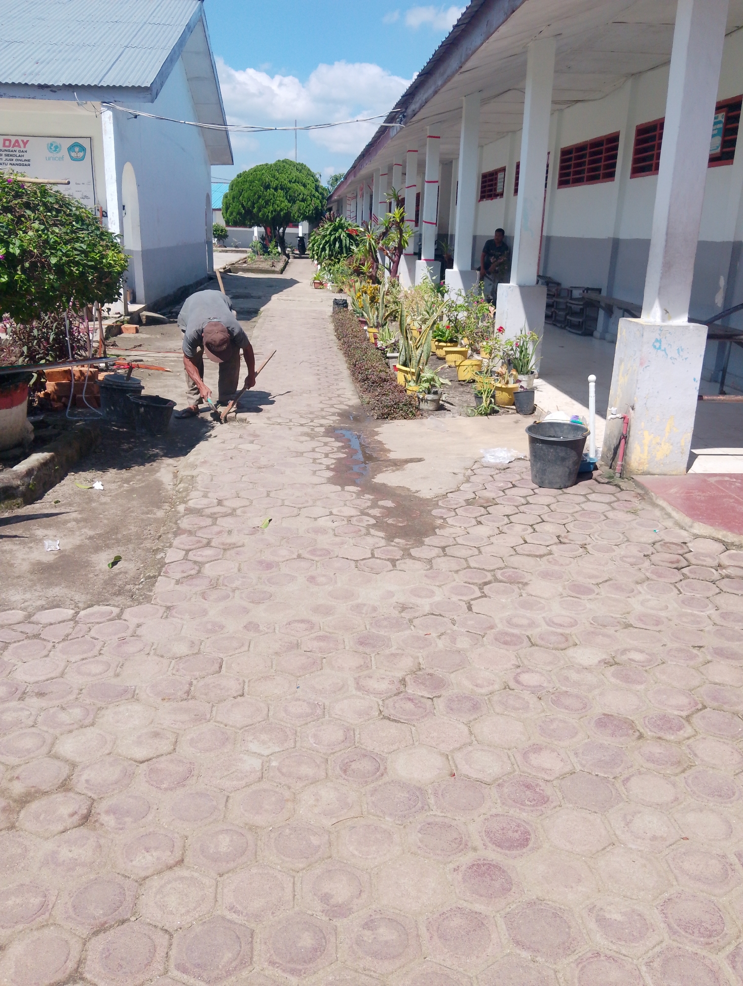 The image depicts an outdoor scene on a sunny day, likely in Serbelawan, Indonesia, showing a walkway paved with hexagonal concrete blocks. In the foreground, a person wearing a grey shirt and a brown cap is crouched down, appearing to be working on the walkway with a tool. Adjacent to the walkway are various potted plants and some construction materials, including bricks.
In the background, a long, white building with red-framed windows runs along the right side of the walkway. The building has a covered porch supported by white pillars. Further down the walkway, another person is partially visible, leaning against the building. On the left side of the image, another white building with a corrugated metal roof is visible, along with a large green bush. The sky is blue with scattered white clouds. The environment appears to be a public space, possibly a school or institutional grounds, given the presence of signage on the left-hand building.