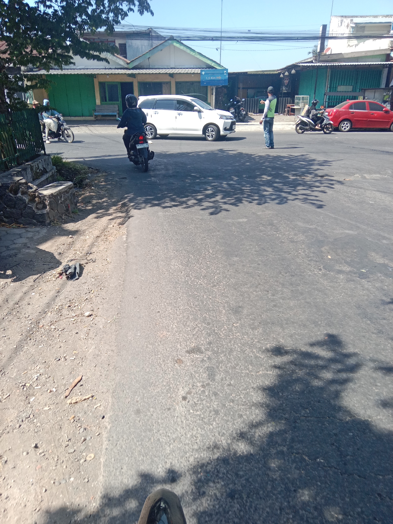 In Dangkel, Indonesia, on a clear, sunny day, a street scene unfolds with a moderate amount of traffic and pedestrian activity. The perspective is from street level, possibly from a moving vehicle.
In the foreground, the asphalt road is dry and shows some signs of wear and tear. The right side of the road is predominantly in shadow from overhead foliage, creating dappled patterns. The left side of the road is lighter in tone and contains scattered debris. A dark, partially obscured object, possibly a piece of cloth or plastic, lies on the ground near a low stone wall.
Further down the road, several vehicles are visible. A white minivan is stopped in the lane. To the right of the minivan, a person in a bright green vest and blue jeans stands, appearing to direct traffic. This individual is facing towards the right of the frame and has their left arm raised. To the right of this person, a red sedan is driving away from the camera. Behind the minivan, a person on a black motorcycle is riding away from the camera, wearing a dark helmet and jacket. Another motorcycle is parked on the side of the road behind the white minivan.
In the mid-ground, a row of buildings lines the left side of the street. These buildings have various colored facades, including green and yellow, with corrugated metal roofs. Some appear to be shops or small businesses. A few trees with green foliage are present, casting shadows on the road and buildings.
On the far left of the image, a green metal fence is visible, bordering the roadside.
The sky is a clear, bright blue, indicating a cloudless day. The overall impression is a typical street scene in a populated area, with daily activities in progress.