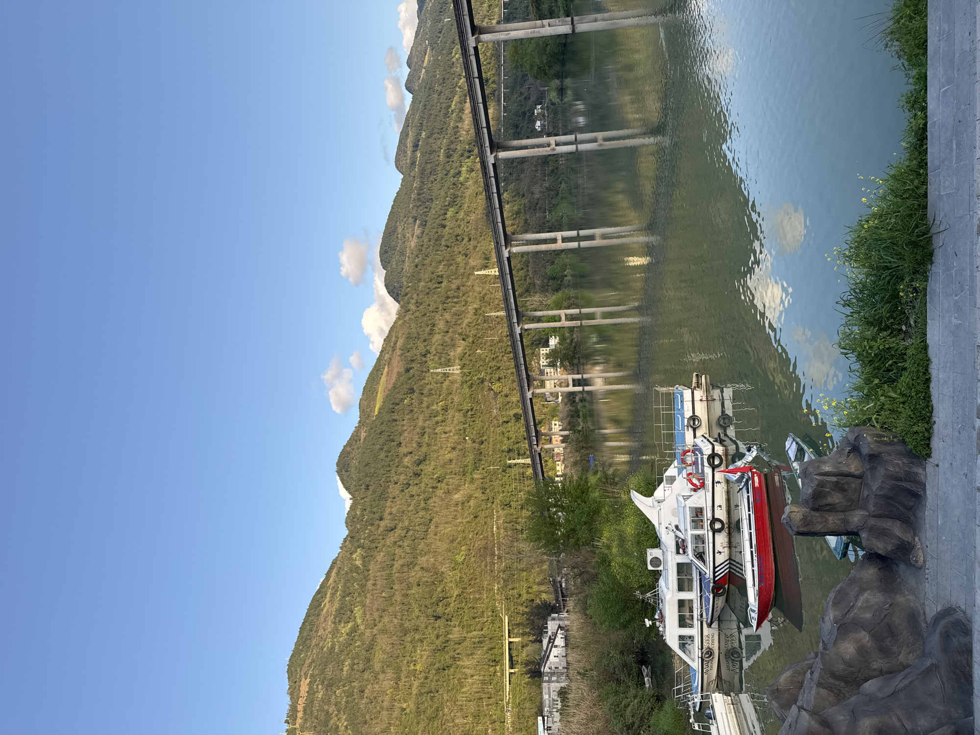 An eye-level, outdoor shot depicts a waterscape scene in Zhuoshui, China, featuring mountains, a bridge, a boat, and the sky. A series of green, tree-covered mountains spans the background under a partly cloudy, blue sky. Reflected in the body of water is a bridge, supported by a series of concrete pillars, running across the water. On the edge of the water, a white and red boat is docked beside a stony area with green vegetation.