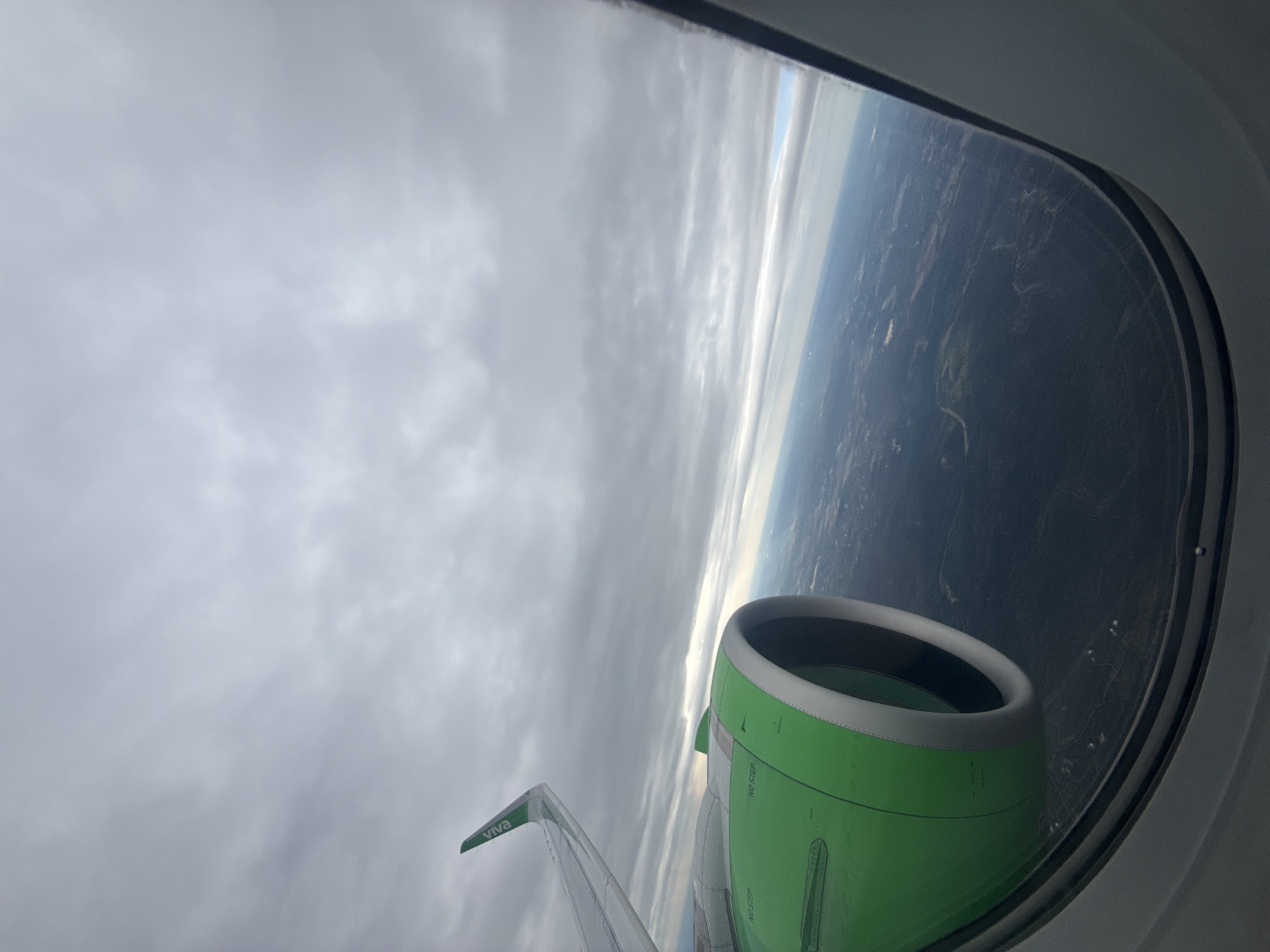 A view from an airplane window reveals a cloudy sky, the wing of the aircraft with the word "VIVA" printed on the tip, and part of the green engine. Outside the window, land is visible, appearing to be a mix of trees and buildings. The location cannot be determined to be San Antonio, United States.