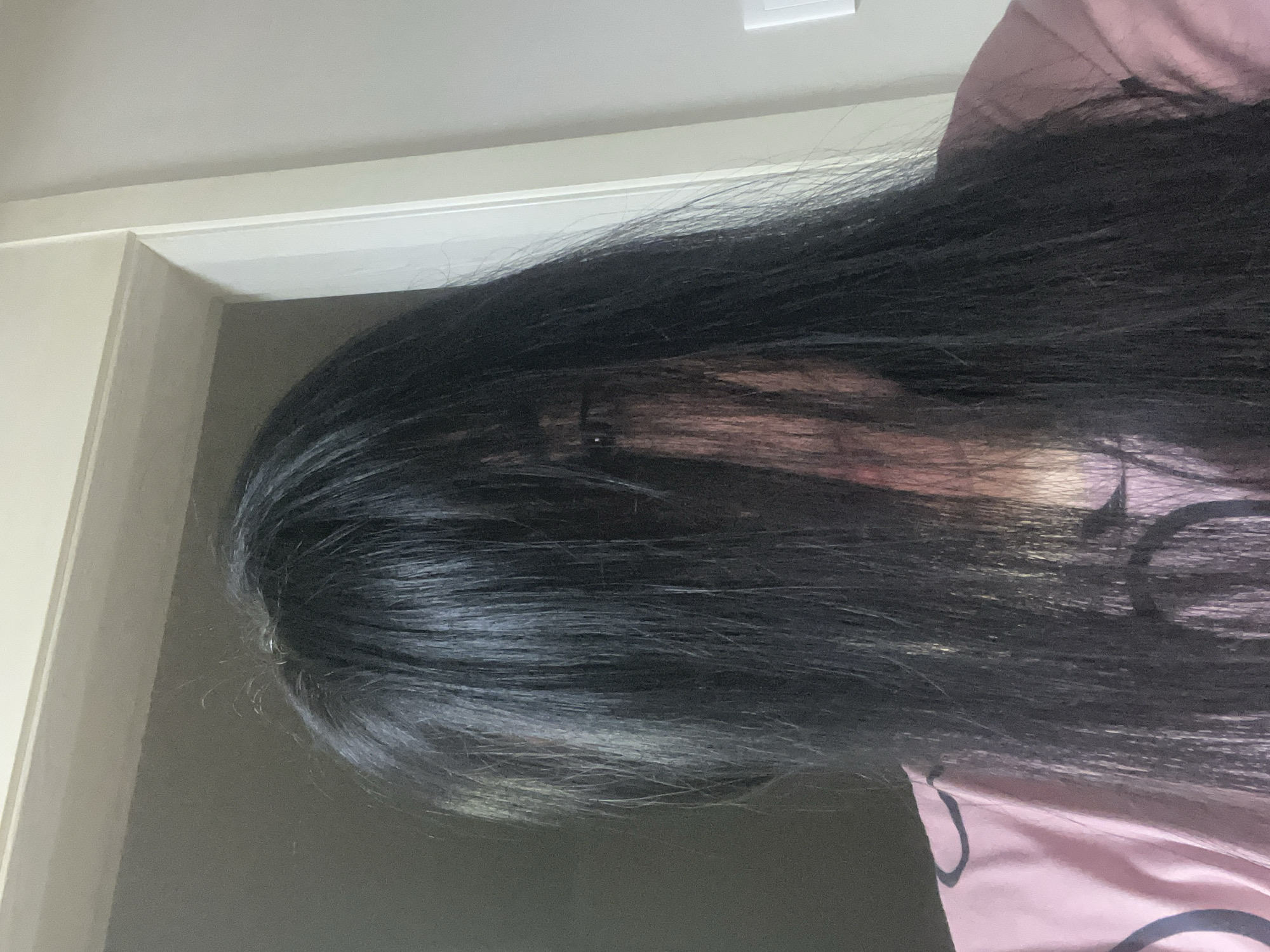 A vertically oriented image depicts a person whose face is almost entirely obscured by a voluminous cascade of long, dark hair. The person's head is positioned on the left side of the frame, with the body extending towards the right. Only a subtle hint of one eye is visible peering through the strands of hair on the upper left side of their face, creating a sense of mystery.
The hair is the most prominent feature, appearing black and flowing, almost acting as a curtain. Some individual hair strands are distinguishable, with a few lighter reflections suggesting depth and texture. The person is wearing a light pink garment, possibly a shirt or top, which is visible along the right and bottom edges of the frame. A faint, dark, curved pattern is noticeable on the pink fabric.
The background consists of a simple indoor setting, with light-colored walls and a door frame visible on the upper left side. The lighting is soft and ambient, providing a gentle illumination without harsh shadows. No specific activity is discernible beyond the person being present with their hair obscuring their face. No text is visible in the image.