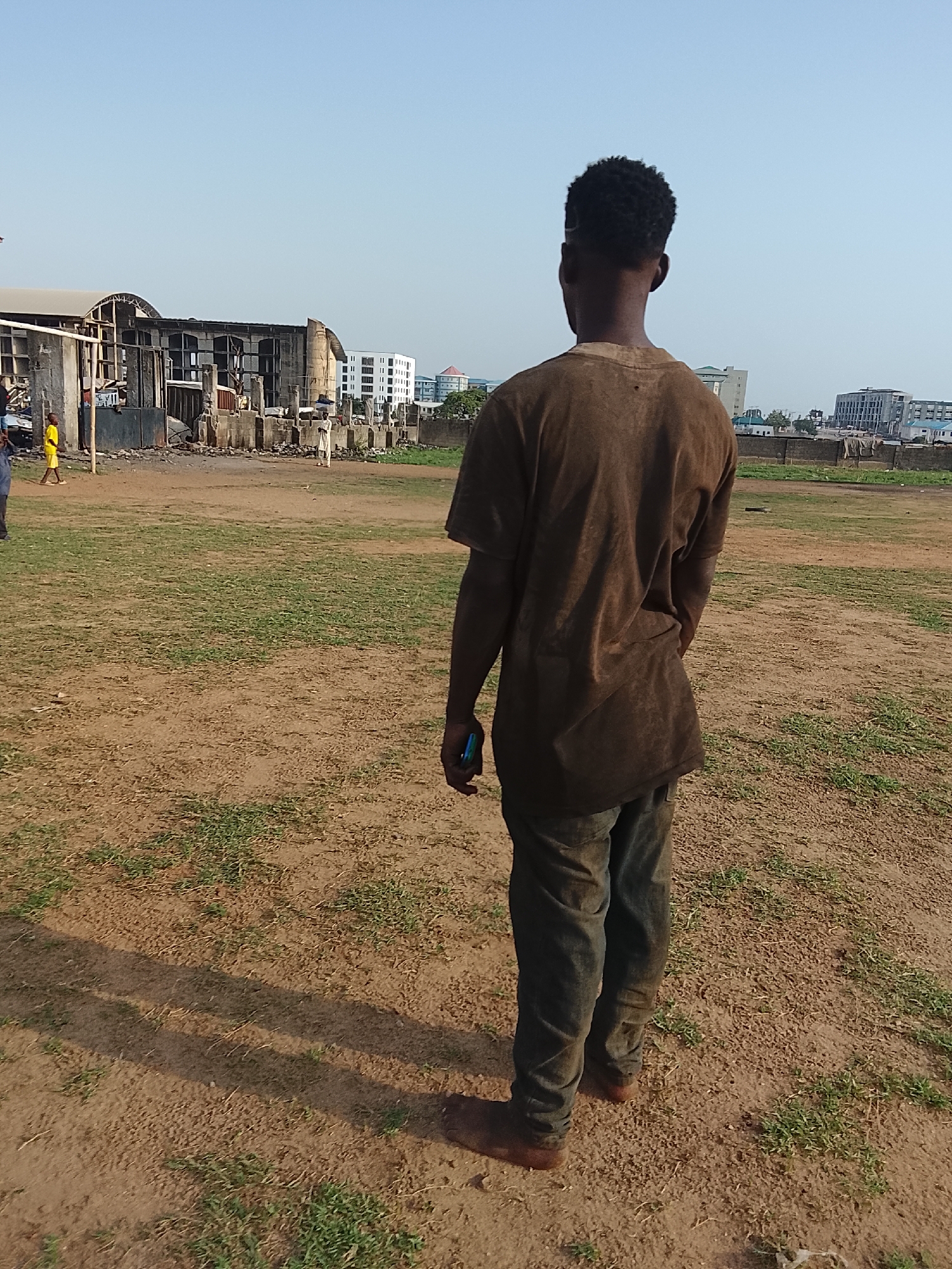 A person is standing on a dirt field with sparse grass. The person is facing away from the camera, wearing a brown shirt and blue jeans. In the background, there is a partially destroyed building, several other people, and buildings in the distance. The location appears to be Abuja, Nigeria.