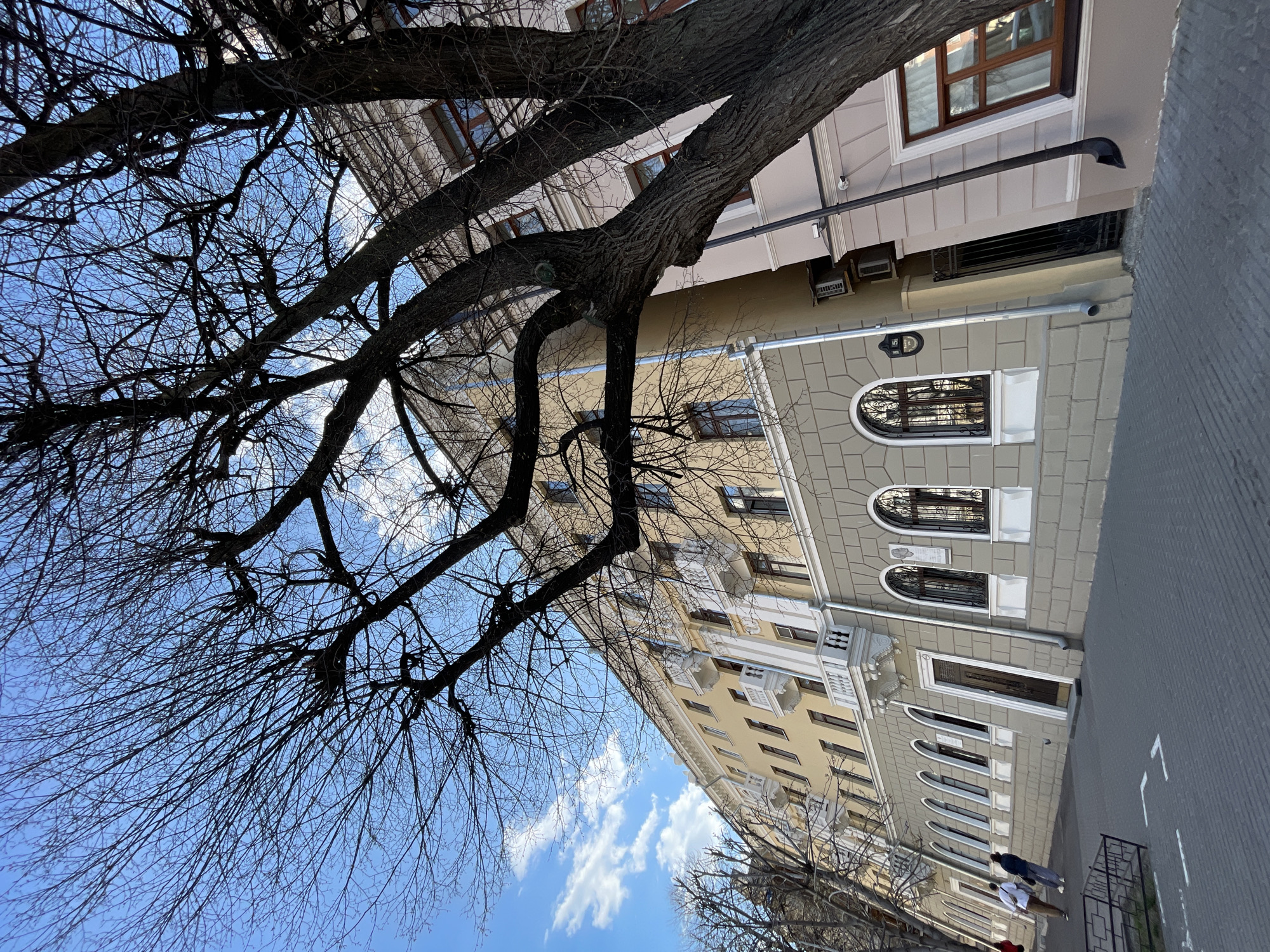 The image depicts a street scene in Kazan, Russia. A multi-story building stands prominently on the right side, featuring architectural details such as arched windows, decorative moldings, and varying shades of beige and gray on its facade. A large tree with bare branches occupies a significant portion of the left side and overhead, partially obscuring the sky, which is a clear blue with scattered white clouds. Two people are visible walking on the sidewalk in front of the building. A metal fence runs along the edge of the sidewalk. The perspective of the photo is angled upward, emphasizing the height of the building and the tree.