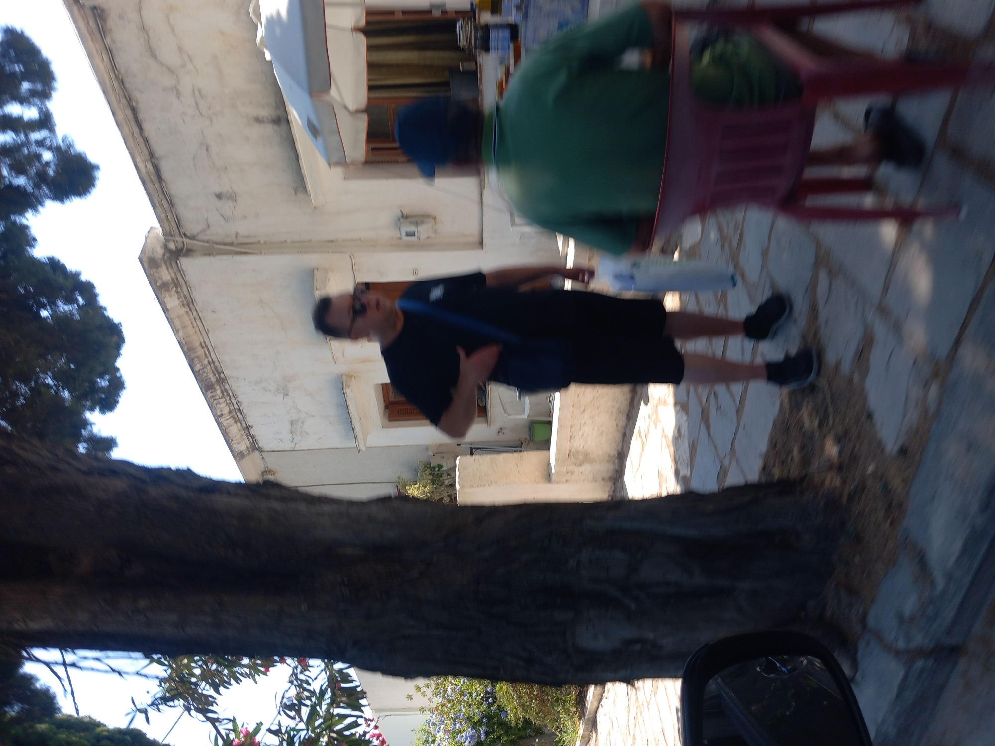 The image captures a street scene in Kos, Greece. A large, rough-barked tree dominates the left side of the frame, its trunk partially obscuring a white building with a tiled roof and visible electrical conduits. In front of the building, a man in a black t-shirt and shorts, wearing sunglasses, stands with a blue bag slung over his shoulder. To his right, another man, seen from the back, wears a green t-shirt and a blue baseball cap, sitting in a red plastic chair. The ground is paved with irregular stone tiles, and shadows suggest a sunny day. A car's side mirror is visible in the lower left corner, indicating the photo was taken from a vehicle. The overall impression is a casual, sun-drenched outdoor setting.