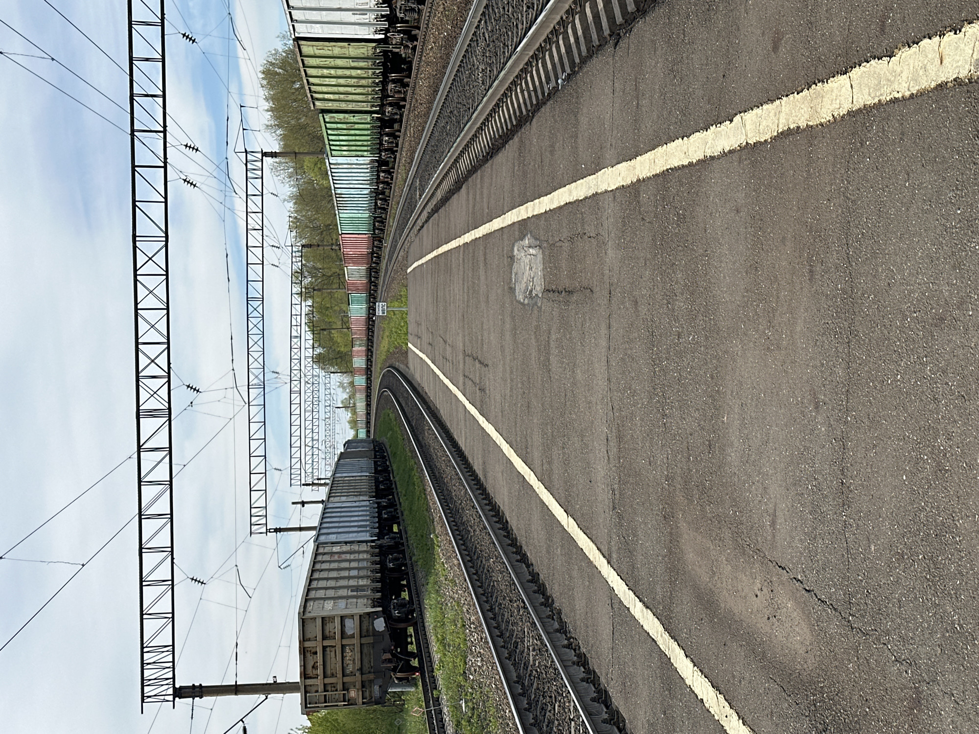 The image depicts a railway platform in Кечёво, Russia. Two sets of train tracks curve through the frame. On the left of the platform, a train with multiple cargo cars is parked, while on the right, a solid white line marks the edge of the platform. Overhead, power lines are suspended by a series of metal towers. The platform itself is paved with asphalt.