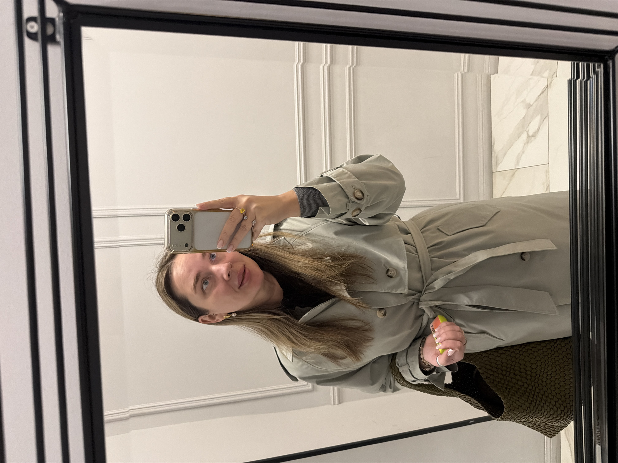 The image is a mirror selfie of a woman indoors, likely in a bathroom or dressing room. She is holding a cell phone in her right hand, taking the picture. The background includes white walls with decorative molding on the left and white marble tiles on the right. The location may be Moscow, Russia, but that is not clear from the picture. She is wearing a light-colored trench coat with a tied belt, and a black top with grey sleeves beneath. She has light brown hair, light skin, and is wearing gold rings on her fingers. A green, woven handbag is visible beneath the coat. The mirror has a thick, black frame with vertical lines.