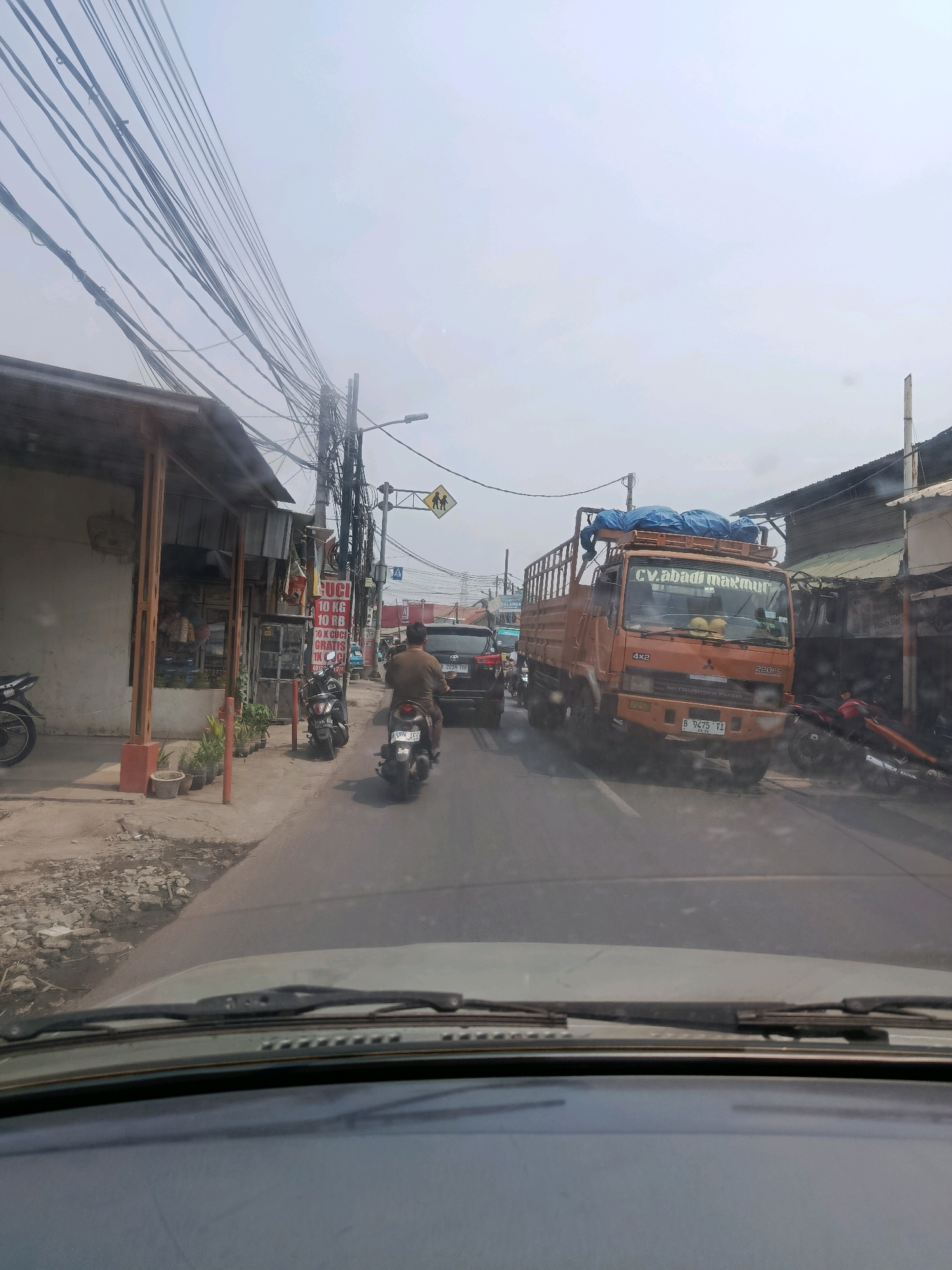The image depicts a street scene in East Jakarta, Indonesia, on a hazy, overcast day. The foreground shows the dashboard and windshield wipers of a vehicle, suggesting the photo was taken from inside a car. The road is lined with buildings and a dense network of overhead electrical wires.
On the left side of the road, there is a simple building with a covered outdoor area, possibly a shop or a small business. A motorcycle is parked to the left of this building. Further down the sidewalk, several potted plants are visible, and a few more motorcycles are parked. A sign with text in Indonesian, which appears to be related to laundry services, is also present.
The road itself is busy with traffic. A man on a motorcycle is in the middle lane, heading away from the viewer. A dark-colored SUV is behind him, and a large orange truck with "CV. ABADI MAKMUR" written on its side is in the right lane. The truck is loaded with blue tarpaulin-covered items. A traffic sign indicating a pedestrian crossing is visible in the background, and another sign with a yellow diamond shape and figures of children suggests a school zone. Various other vehicles and structures are present in the distance, indicating a typical urban environment. The overall atmosphere is that of a moderately congested street in a commercial or residential area of the city.