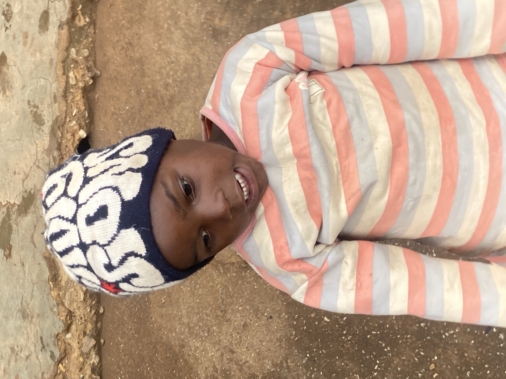 A young child, appearing to be of African descent, stands outdoors wearing a white knit beanie with blue lettering and red accents. The child is also wearing a long-sleeved striped shirt with alternating bands of pink, white, and light blue. The child is looking down and to the right, with their mouth slightly open. The background consists of a textured, earth-toned wall and ground, suggesting an outdoor urban environment in Bukuru, Nigeria.