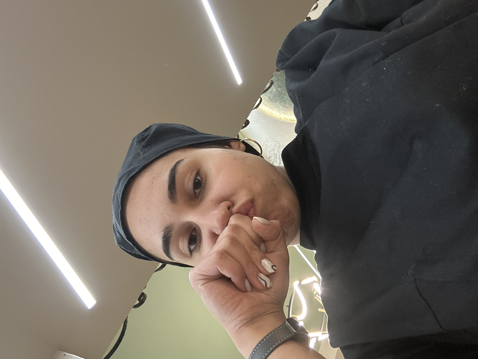 A woman with tan skin and dark eyebrows and eyes looks directly at the camera with a slight pout, her hand covering part of her mouth. She wears a black head covering and a black hooded sweatshirt. A gray watch is visible on her left wrist. The background includes a light-colored ceiling with a long, linear light fixture and a green wall with decorative elements. While a location is mentioned in the prompt, Burgas, Bulgaria, the image does not contain enough information to determine the exact location.
