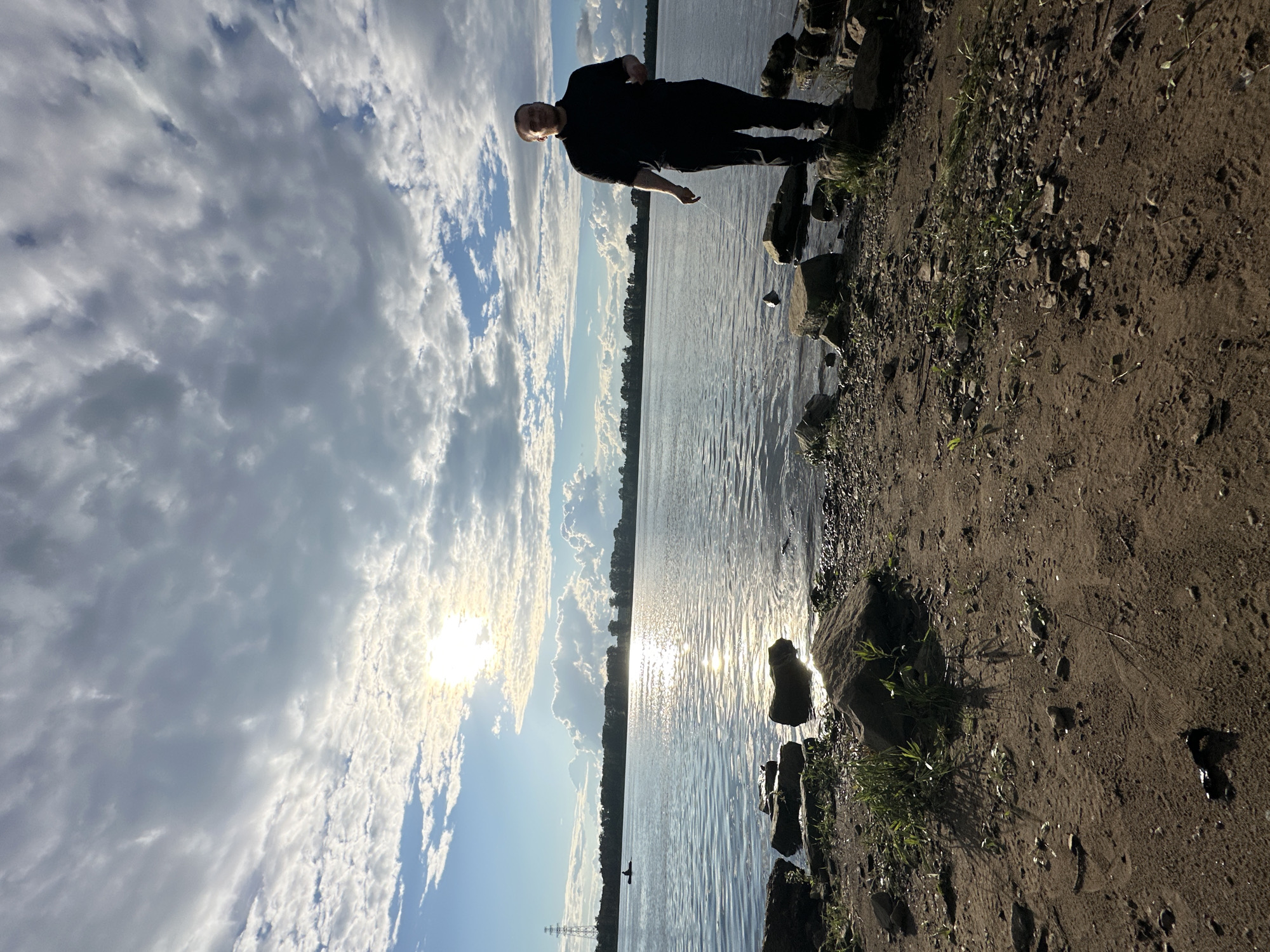 A person stands on a rocky shore overlooking a body of water, possibly a river or lake, with a line of trees in the distance. The sun is low in the sky, casting a warm glow and reflecting on the water's surface. The sky is filled with a mix of fluffy white clouds and darker, more dramatic clouds, suggesting a partly cloudy day with potential for changing weather. The person is dressed in dark clothing and appears to be looking out at the water or the horizon. The ground is composed of dirt, pebbles, and some sparse vegetation. There is a power transmission tower visible on the far shore, indicating proximity to human infrastructure. The overall mood is serene and contemplative, with the natural beauty of the landscape taking center stage. The time of day appears to be late afternoon or early evening, given the angle of the sun. The location is identified as Chaikovsky, Russia.