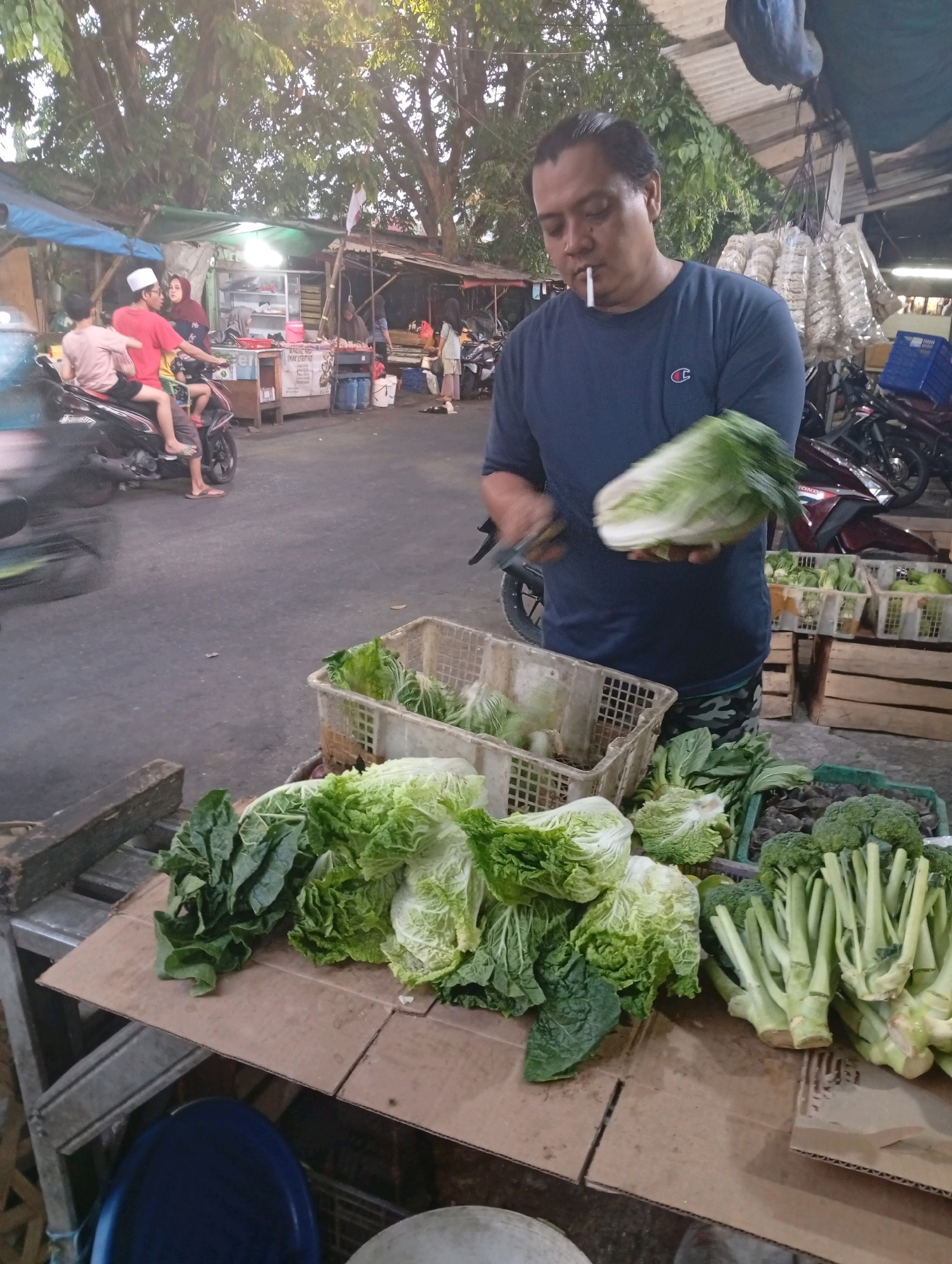 A man in a blue t-shirt holding a head of napa cabbage and a cigarette stands in front of a vegetable stall. He is in a street market environment, possibly in North Jakarta, Indonesia. The stall displays various vegetables like napa cabbage, broccoli, and leafy greens in baskets and crates. In the background, a motorcycle with two people is visible, and further back, there are more stalls and trees. The scene suggests a typical market setting with goods and people engaged in commerce.