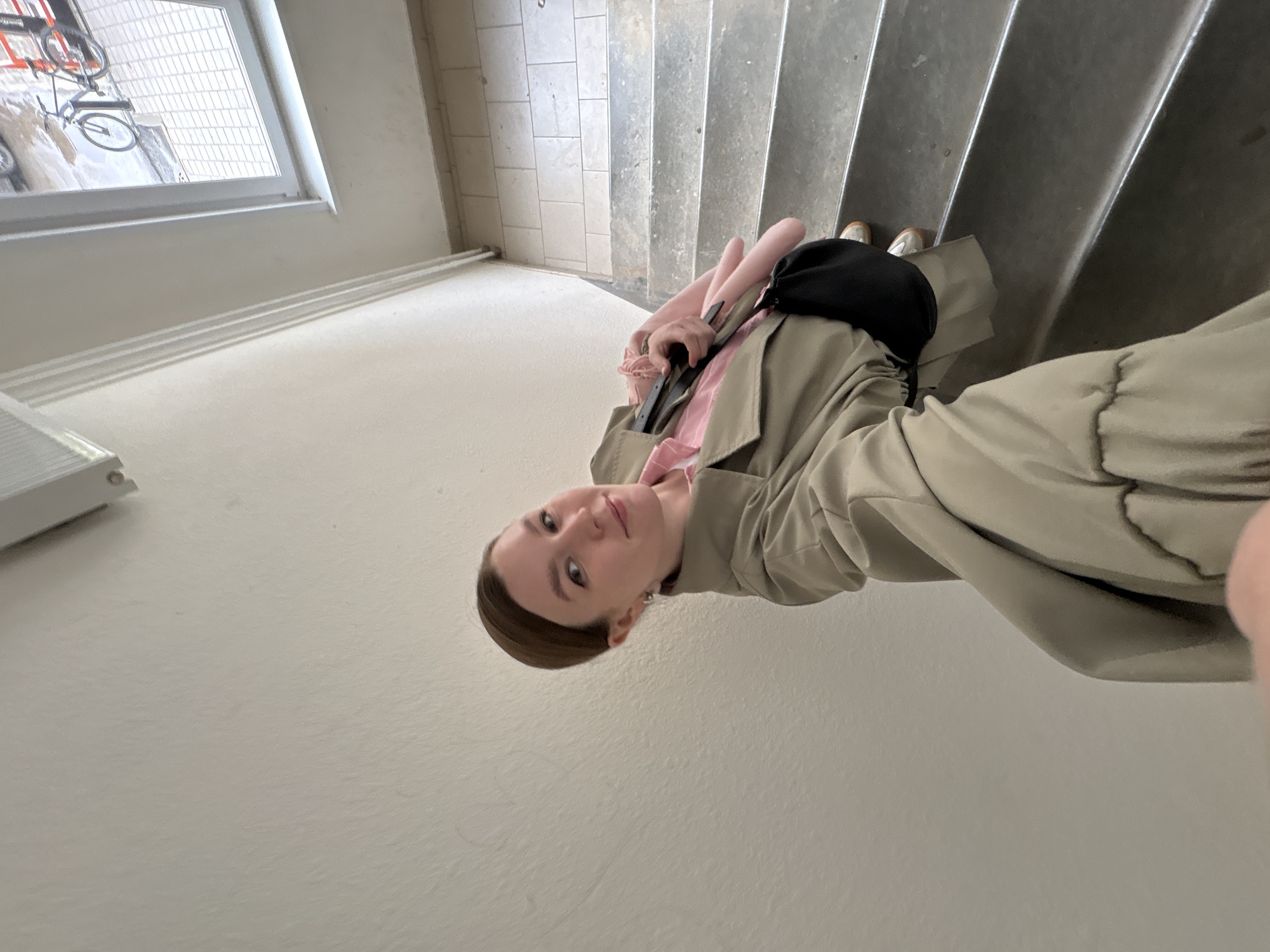 A young woman is captured in a selfie taken indoors on a staircase, in what appears to be Izhevsk, Russia. She is positioned on a concrete staircase with tiled landings visible on the left. The image is rotated, showing her looking directly at the camera with a neutral to slightly pleasant expression.
The woman has light brown hair pulled back and is wearing a light beige or khaki trench coat over a pink and white striped shirt. She carries a black shoulder bag on her left side and holds a pink, rolled item, possibly a yoga mat or blanket, along with a dark strap in her right hand. Her feet, clad in light-colored shoes, are visible on the step below her.
In the background, a large window reveals an outdoor scene. Through the window, a bicycle with thin tires is visible, along with what appears to be a paved area covered with snow or ice, suggesting cold weather conditions outside. The interior walls are plain and light-colored, and white pipes or railings run along one wall beneath the window. No discernible text is visible in the image. The overall setting is utilitarian, likely a residential building or public stairwell.