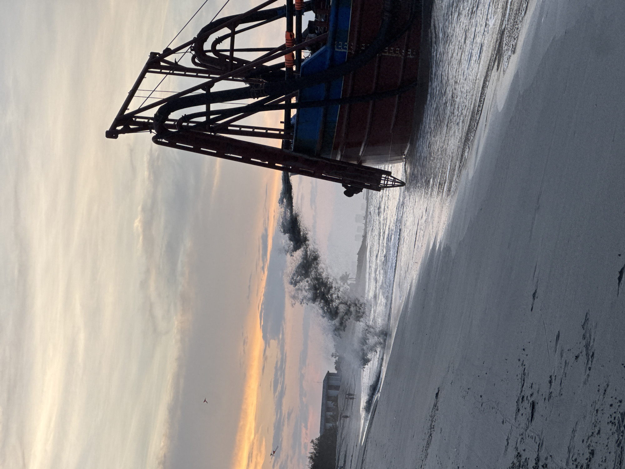 A fishing trawler is beached on a sandy shore. The boat is rusted orange and blue. The boat's front end is facing forward. The boom of the boat is visible. The ocean is visible on the right of the picture. The sky is overcast with yellow, white, and gray colors. People and buildings are in the mid-ground on the bottom right. This appears to be My Khe beach in Đà Nẵng, Vietnam.