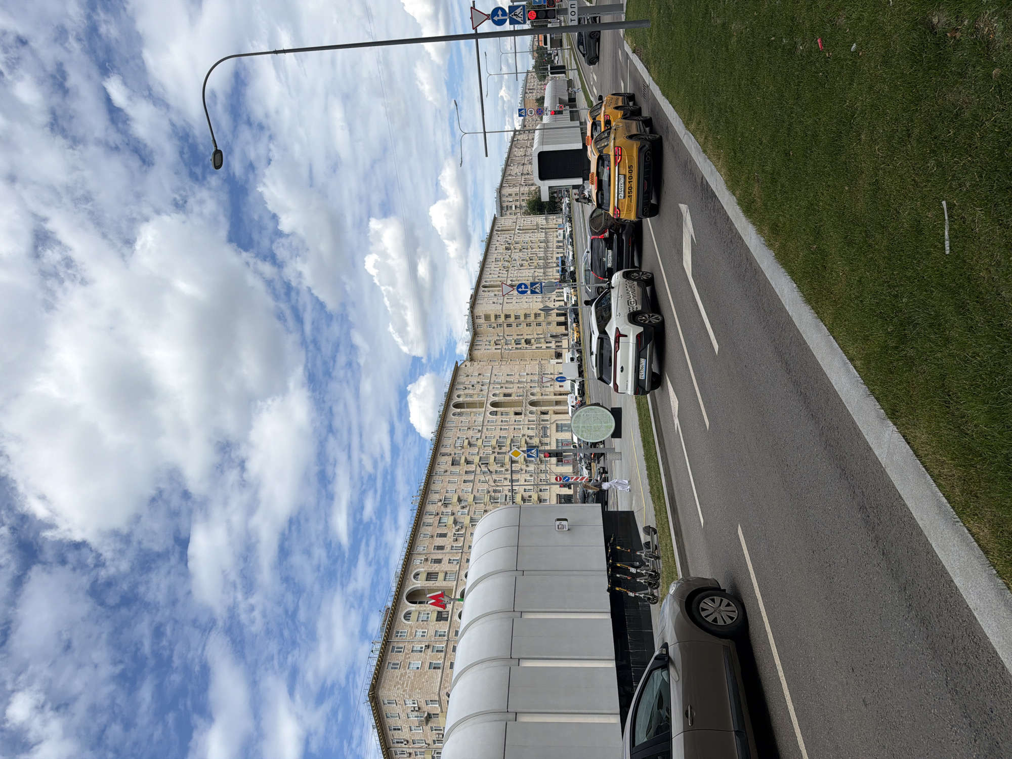 An eye-level, outdoor shot captures a street scene in an urban environment under a partly cloudy sky. The focal point is a wide asphalt road with white lane markings, carrying a moderate flow of traffic. Several cars, including white SUVs and yellow taxis, are visible on the road, some of which are stopped at a traffic light. On the right side of the road, a patch of vibrant green grass borders the asphalt, with a few scattered pieces of debris. A tall street lamp with a curved arm stands on the right edge of the frame.
To the left of the road, a wide sidewalk features a long, modern, arched structure, likely part of a public transportation station, painted in a light gray hue. Several electric scooters are parked in a row next to this structure. Beyond the sidewalk, a row of imposing, multi-story apartment buildings stretches along the street. These buildings are characterized by their light-colored facades, numerous windows, and arched entrances on the ground floors. A prominent red metro sign with a white "M" is visible on one of the buildings, indicating a subway station entrance. The overall impression is a bustling city street with a blend of residential architecture and modern infrastructure.