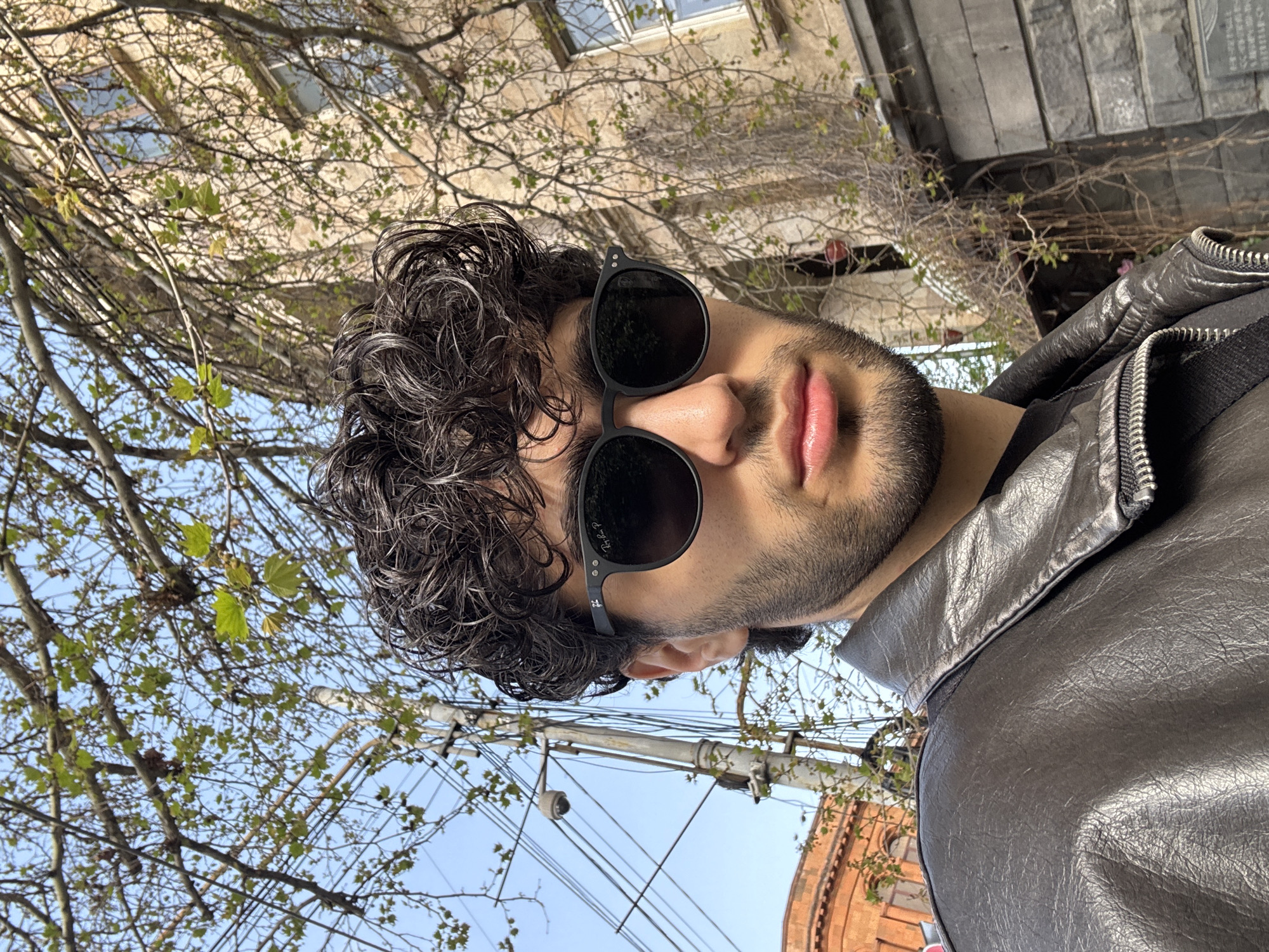 The image shows a man wearing sunglasses and a leather jacket, presumably outdoors. The background includes buildings and trees. Based on the architectural style, the location appears to be Yerevan, Armenia. The buildings are likely stone or concrete, possibly residential or commercial. There are utility poles and wires visible against a clear sky. The man is the primary subject, with the surroundings suggesting an urban environment.
