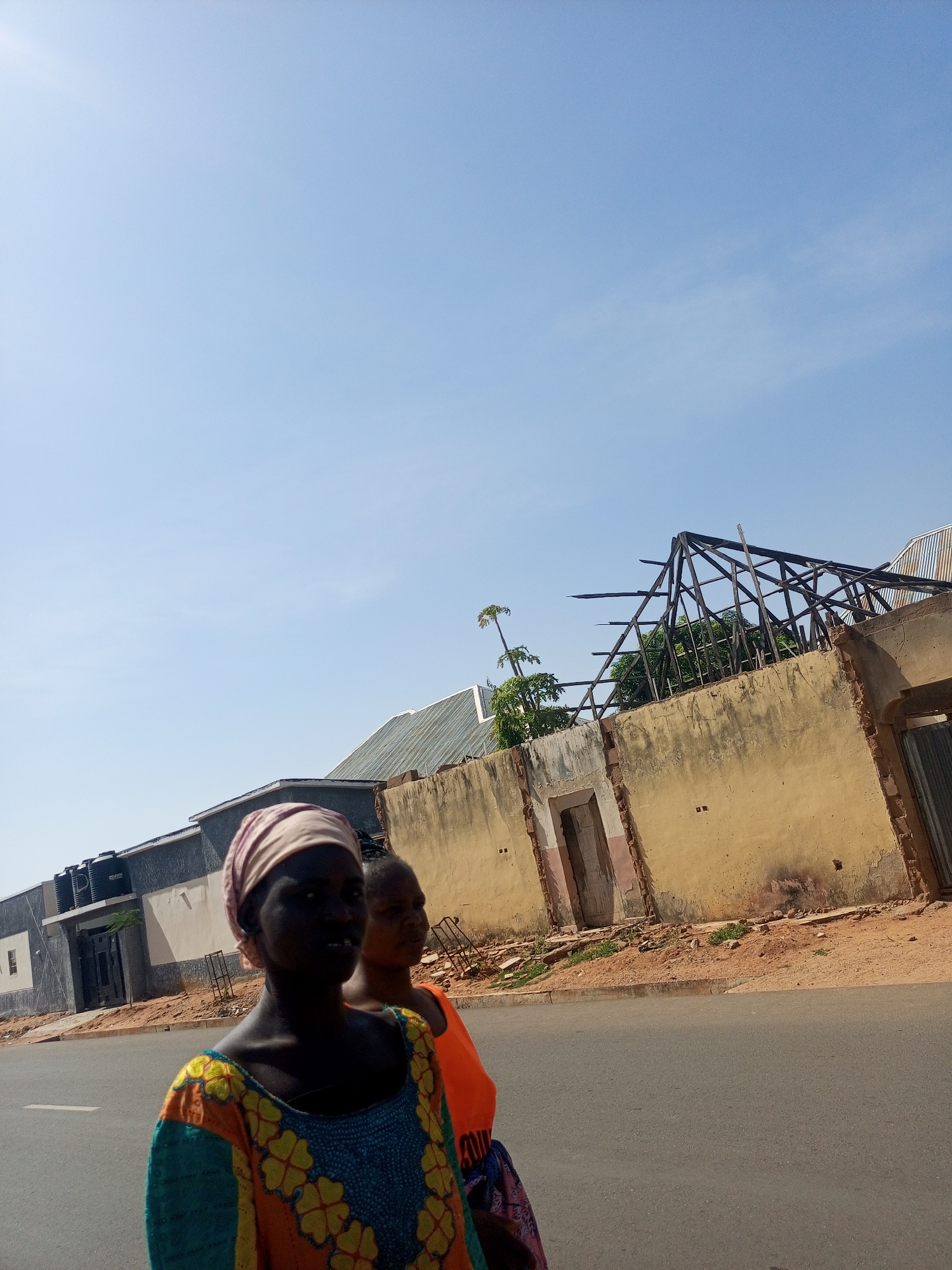 Two women walk down a street in Unknown, Unknown. One woman wears a headscarf and a brightly colored dress. The other woman wears an orange shirt. Behind them are several buildings. One building has a partially collapsed roof.