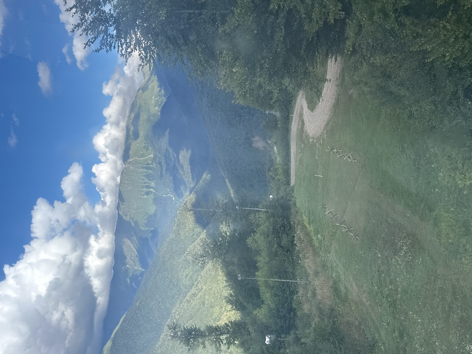 The image depicts a scenic mountain landscape in Estosadok, Russia, on a clear day with fluffy clouds in the sky. The foreground features a grassy slope with a dirt path winding through it. Several tall, thin posts with lights are visible on the left side of the path, along with some sparse vegetation. In the mid-ground, dense green trees form a lush forest. Beyond the trees, majestic mountains rise, their slopes covered in vegetation and dappled with sunlight and shadow. The overall scene conveys a sense of natural beauty and tranquility.