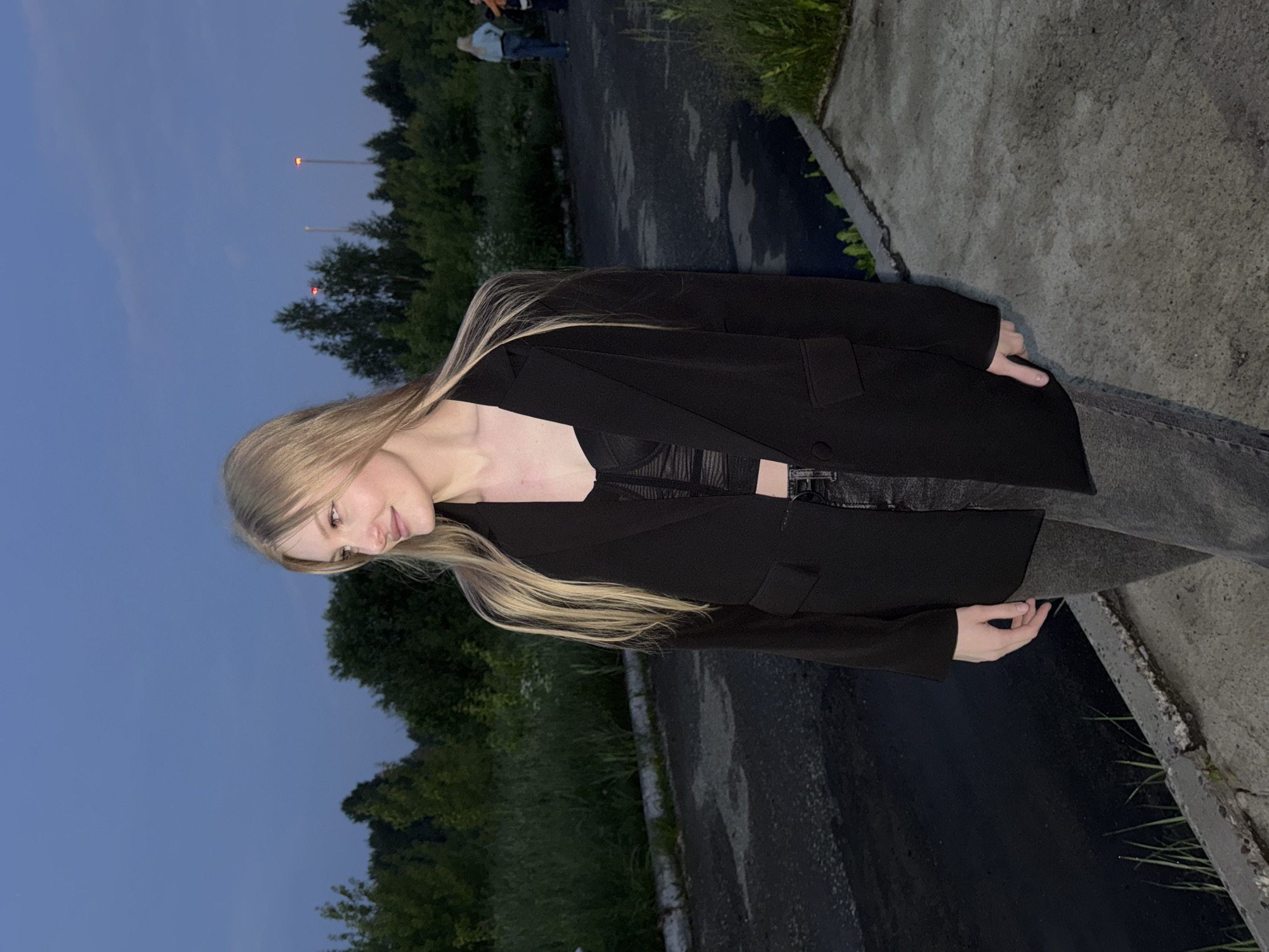 A young Caucasian woman with blonde hair and light skin is standing outdoors at dusk, looking to her left. She is wearing a black blazer over a black lace bralette and dark grey jeans. Her left hand is resting on her hip, and her right arm is slightly bent. The background consists of a paved road with patches of water, a grassy embankment, and a line of trees. In the distance, a few people can be seen, and tall poles with lights are visible against the twilight sky. The scene appears to be on the outskirts of the city of Perm, Russia, possibly near a road or industrial area given the presence of the paved surface, embankment, and distant lights.