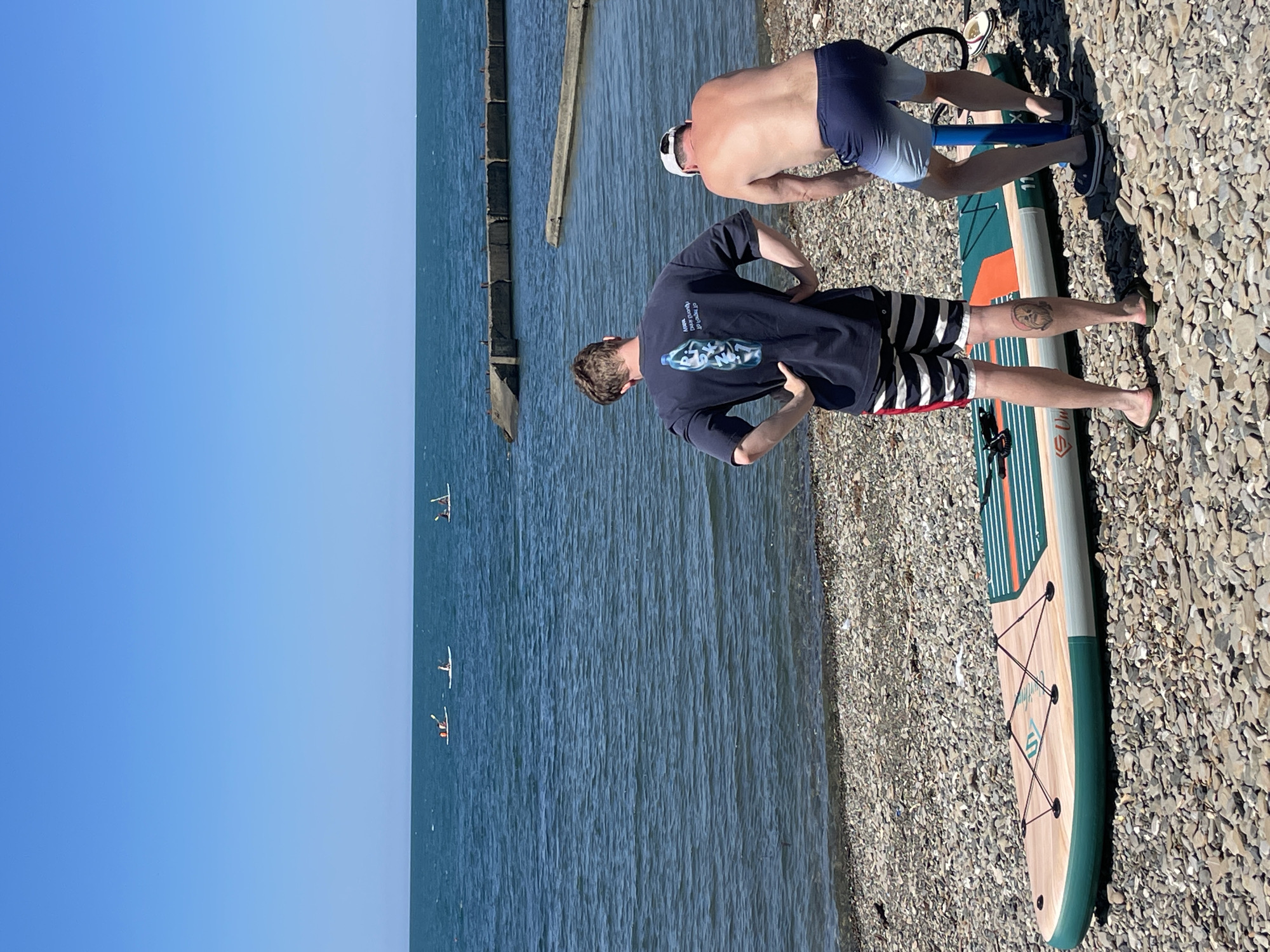 In Khostinskiy, Russia, two individuals prepare for paddleboarding on a sunny day. The scene is set on a pebble beach with the Black Sea in the background. A stand-up paddleboard, partially inflated, lies on the ground. One person, wearing a dark t-shirt and striped shorts, stands with hands on hips, facing the water. The other, shirtless and wearing blue shorts, is operating a manual pump connected to the paddleboard. In the distance, several people are visible on paddleboards in the calm, blue water. The shoreline is lined with concrete structures, likely remnants of a pier or breakwater. The sky is clear and bright blue.