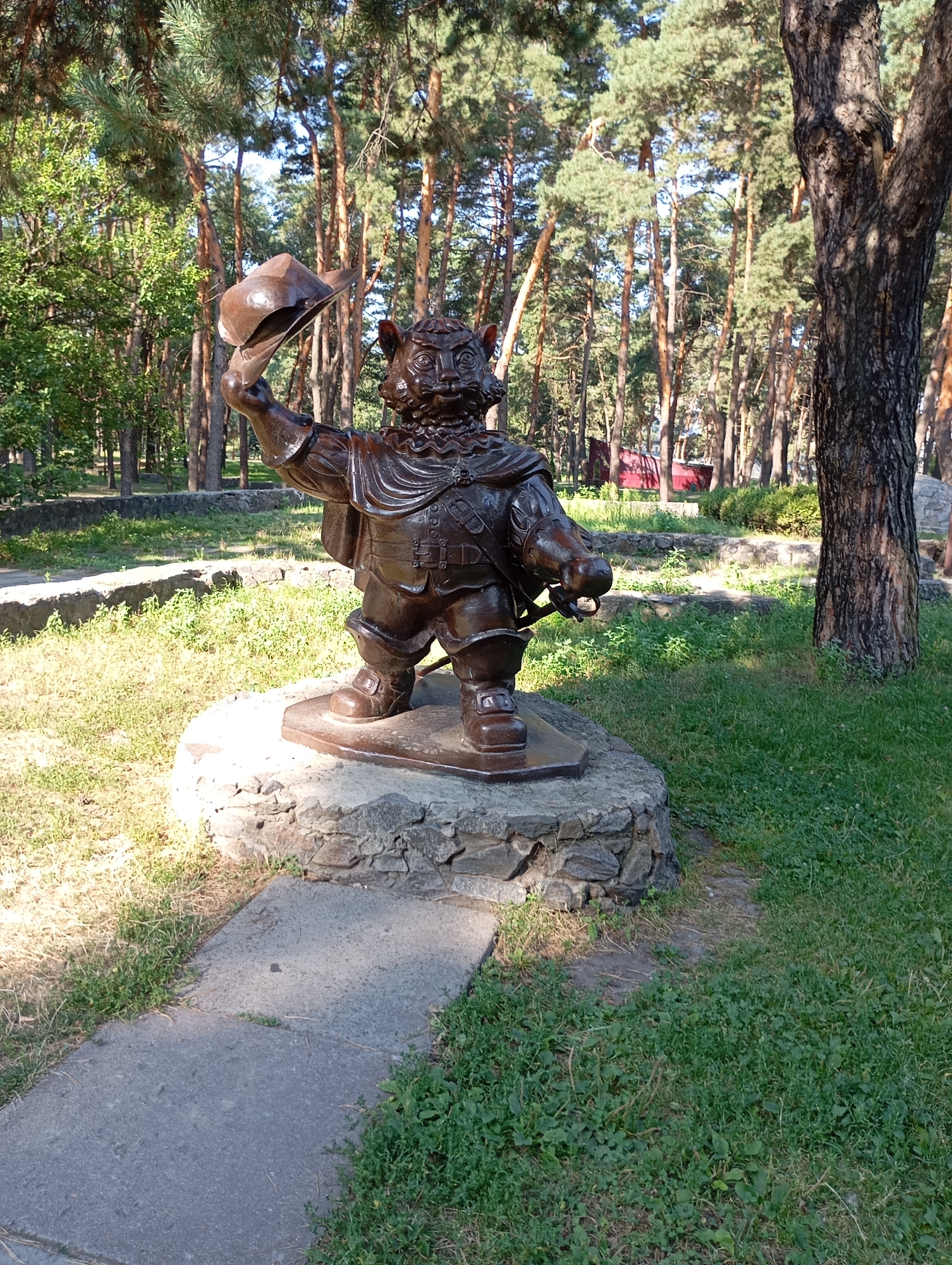A bronze statue of a cat-like character in a hat and cape stands on a stone pedestal in a park. The statue is depicted as a bipedal feline wearing a ruffled collar, a cape secured with clasps, a tunic with a belt and buckle, and boots. The character holds a wide-brimmed hat in its raised left hand, and its right hand is extended forward, holding a sword or similar object. The statue is positioned on a circular stone base, which itself rests on a larger, irregularly shaped stone platform. The surrounding environment is a park with mature trees, primarily pines, with a grassy area and a paved path leading to the left of the statue. In the background, there are hints of other structures and landscaping, including stone retaining walls and what appears to be a red building. This is a public park in the city of Horishni Plavni, Ukraine, specifically in the area known for its recreational spaces and monuments. The lighting suggests a sunny day, casting shadows on the ground and the statue.