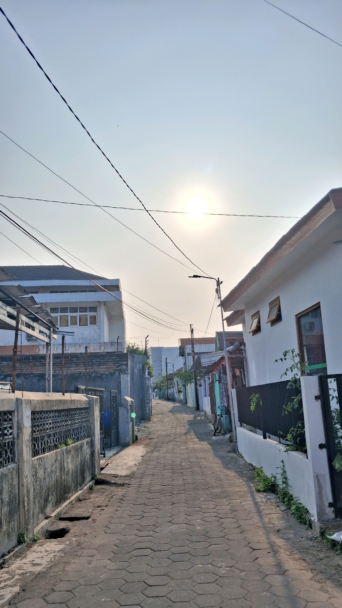 A narrow, paved alleyway lined with houses and buildings stretches into the distance under a bright, hazy sky. The sun is visible, low in the sky, casting a soft glow. Overhead, numerous electrical wires crisscross the frame. On the left, a concrete wall with decorative metalwork borders the alley. Beyond it, a multi-story building with white siding and rows of windows is visible. On the right, smaller, single-story houses with white walls and dark trim line the alley. One house has two small windows propped open. A black metal fence runs along the front of another house, with greenery growing through it. A bicycle is parked further down the alley on the right side. The pavement is made of hexagonal interlocking blocks. The scene suggests a residential area in a city, possibly in Indonesia, given the architectural style and the presence of overhead wires common in many Southeast Asian cities. The overall atmosphere is calm and quiet, likely captured during the early morning or late afternoon.