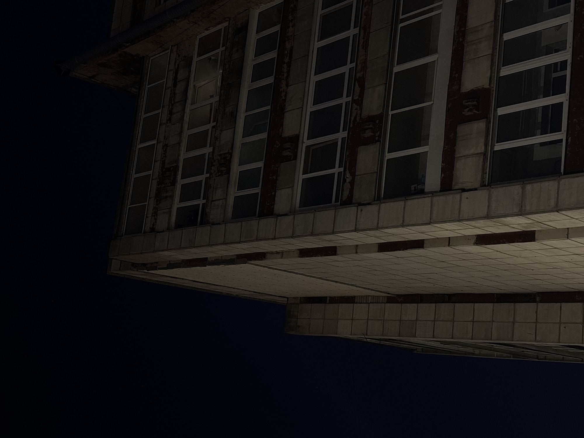 The image shows the exterior of a building with multiple windows. The building is located in Perm, Russia. The perspective is from a low angle, looking up at the side of the building. The sky is dark, suggesting it is night. The building appears to be constructed of concrete and has a tile-like pattern on its underside. The windows are rectangular and divided into smaller panes. The building appears to be old and somewhat dilapidated.