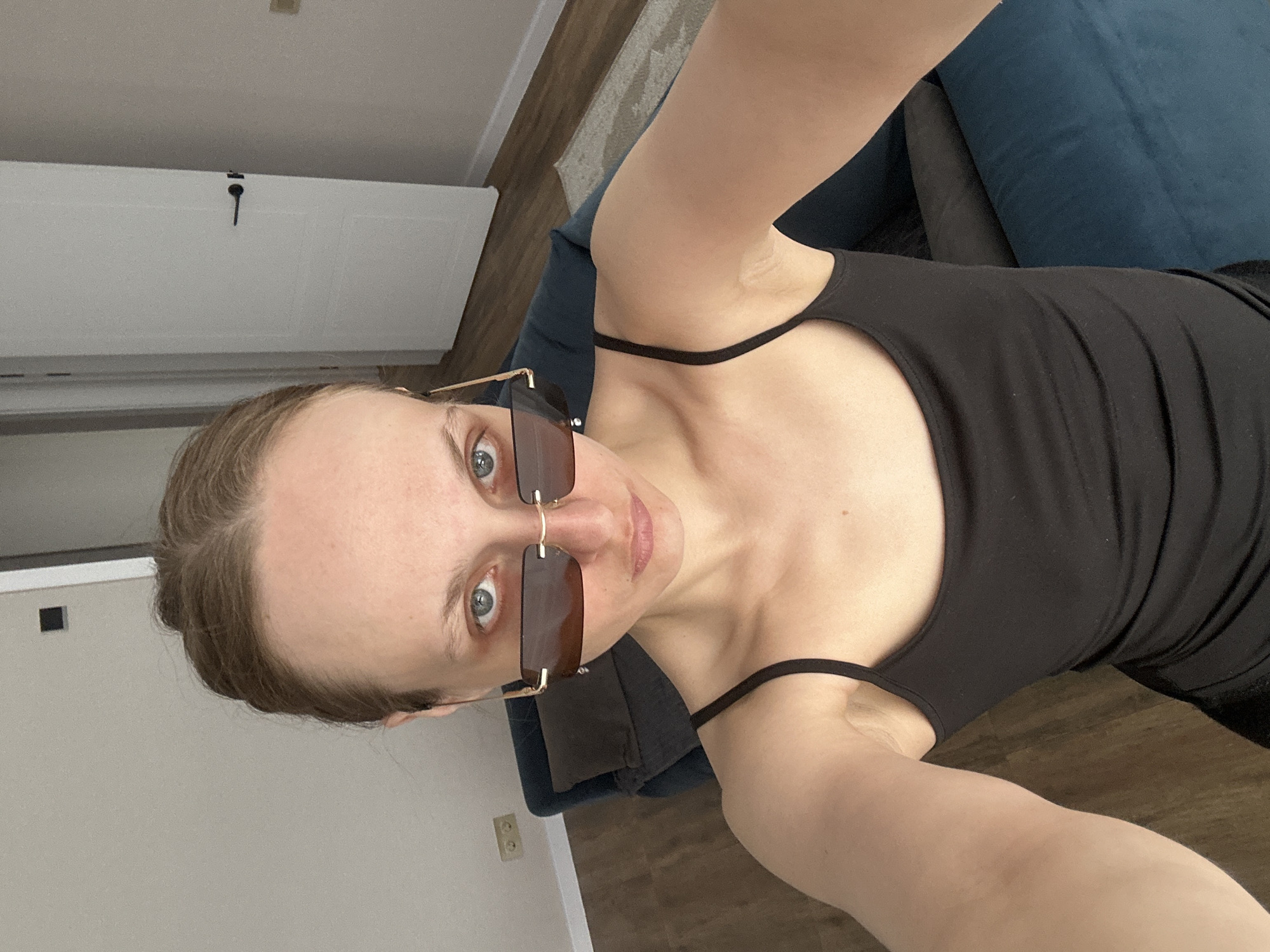 A selfie of a fair-skinned woman with light brown hair pulled back, wearing dark-tinted sunglasses and a black tank top. She is sitting or reclining on a blue upholstered chair. The background includes a white door with a black handle, light gray wall, and a section of wood-look flooring. The location is potentially Sepych, Russia, based on the user's location, but there are no distinct landmarks visible in the image to verify this. The interaction consists of the woman taking a selfie.