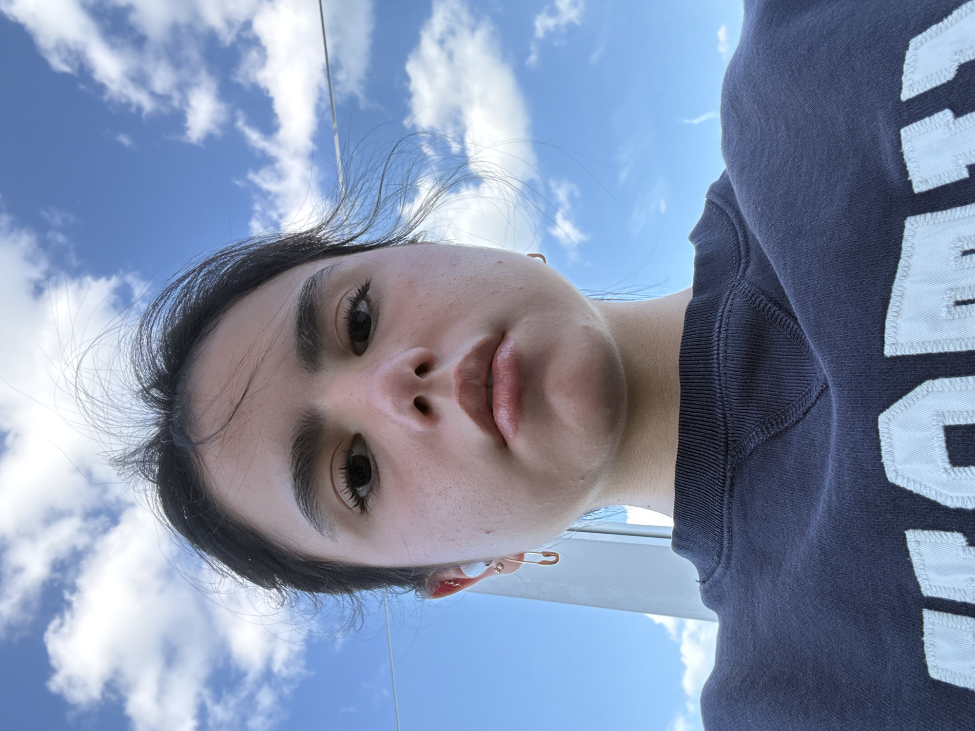 The image is a close-up selfie taken from a low angle, facing upward towards the subject's face and the sky. The subject appears to be a young woman with dark hair and light skin. The background consists of a bright blue sky with scattered white clouds. A portion of a dark blue sweater with white lettering is visible on the right side of the frame. The subject is wearing a gold safety pin earring. The location cannot be determined from the image.